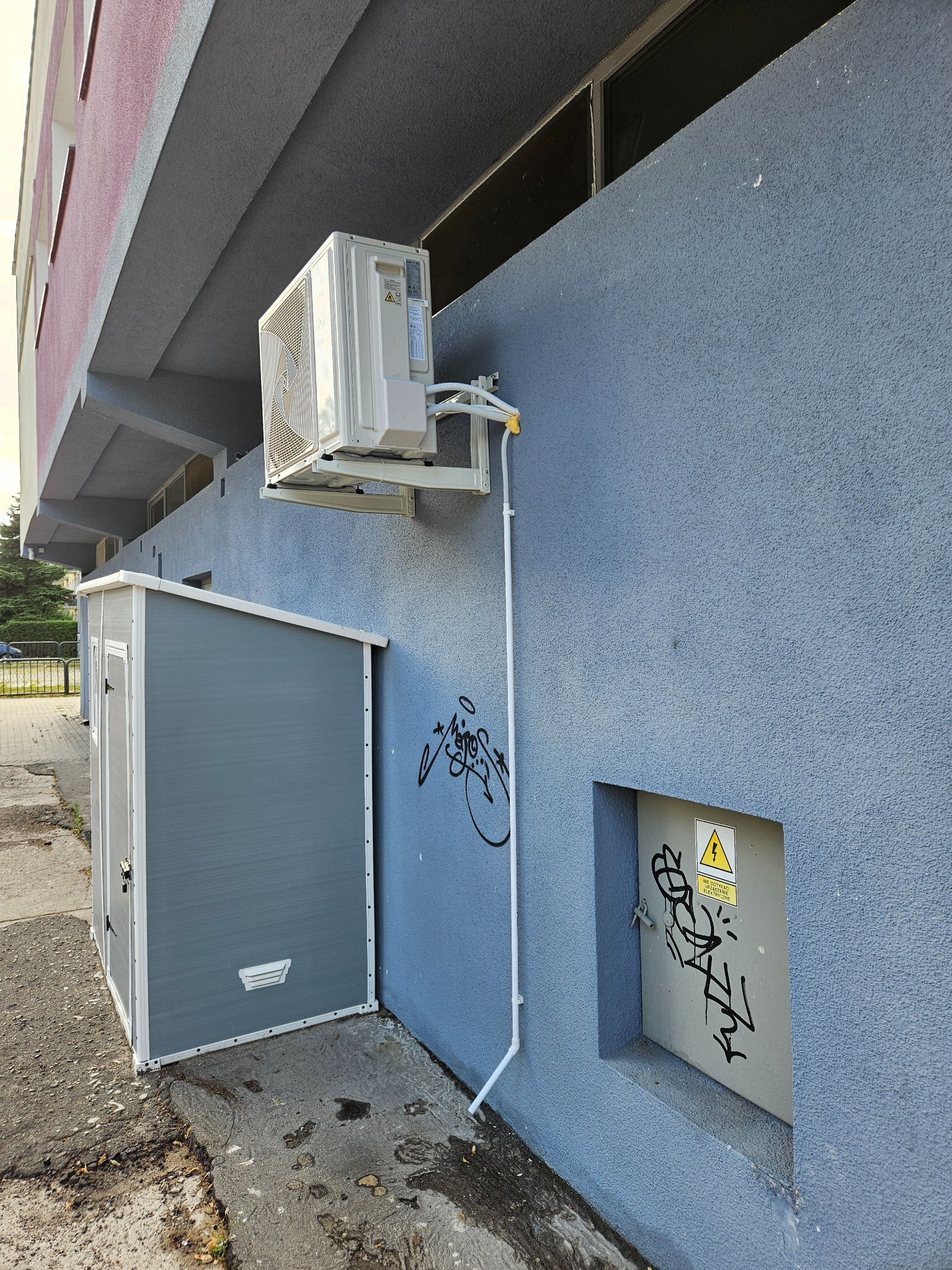 Biała jednostka zewnętrzna klimatyzatora zamontowana na niebieskiej ścianie budynku, obok szara szafka narzędziowa i skrzynka elektryczna z ostrzeżeniem i graffiti.