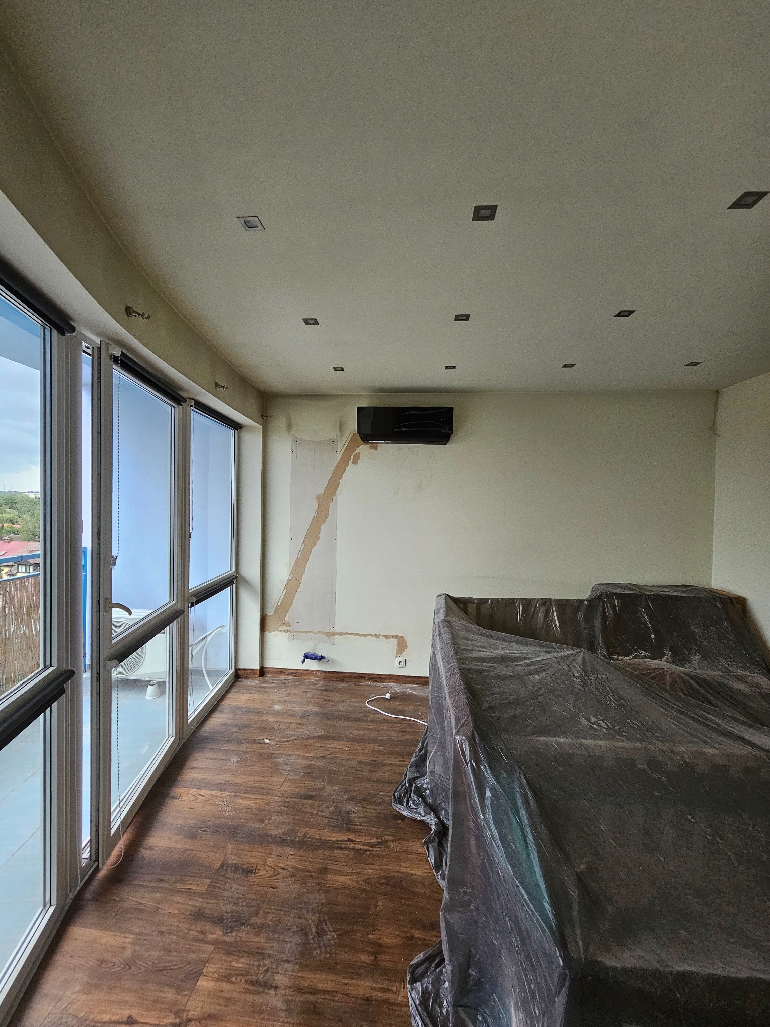 Wnętrze pokoju z zamontowanym czarnym klimatyzatorem ściennym, widoczne przygotowania do prac wykończeniowych: meble zakryte folią ochronną, ślady po szpachlowaniu na ścianie, podłoga wyłożona...