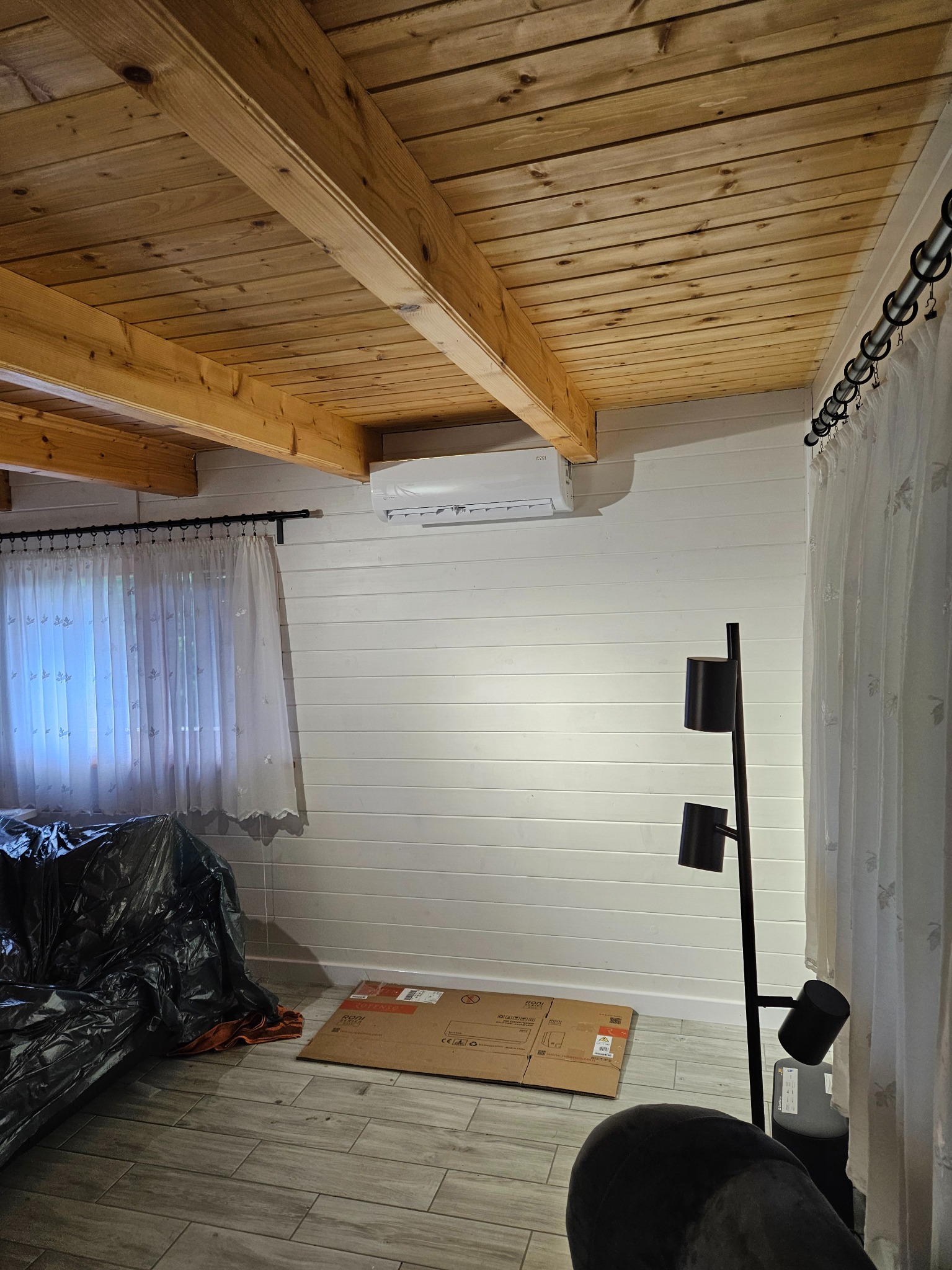 Wnętrze pokoju z drewnianym sufitem i zamontowanym klimatyzatorem na białej ścianie obitej boazerią; widoczny karton na podłodze i zasłonięty mebel.