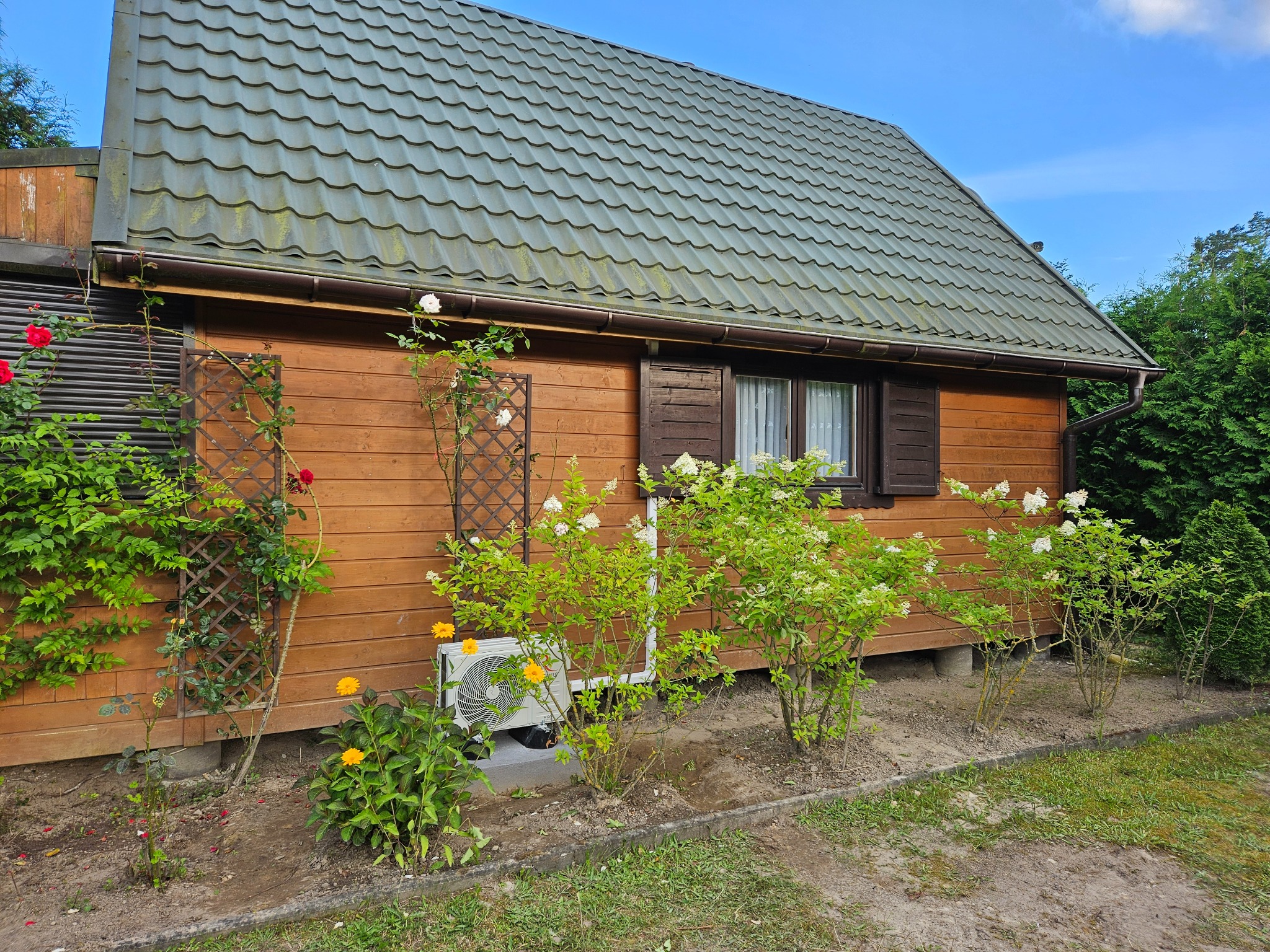 Zewnętrzna jednostka klimatyzatora zamontowana na betonowej podstawie przy drewnianym domku, otoczona kwitnącymi krzewami i kwiatami w ogrodzie.