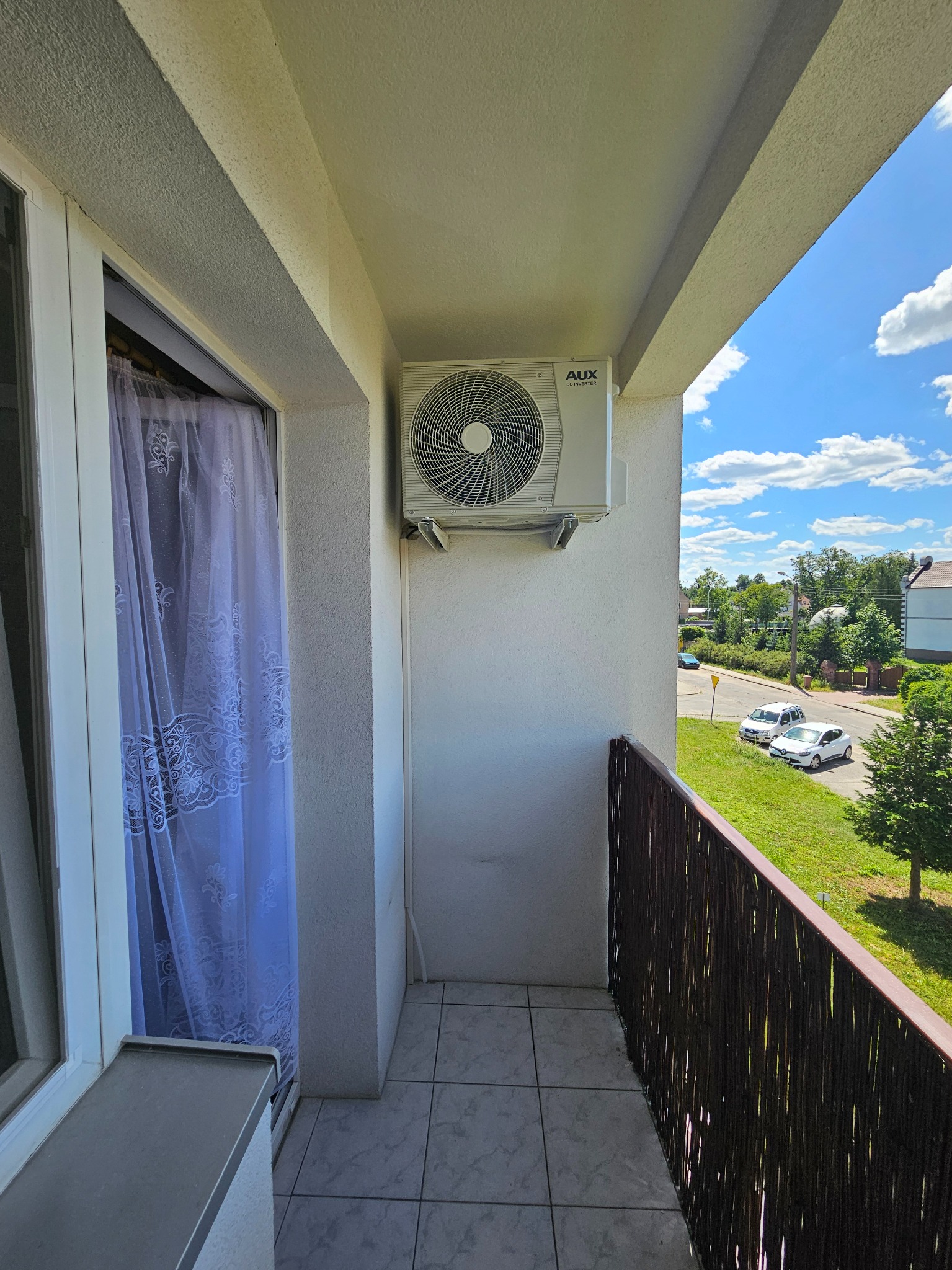 Klimatyzator AUX zamontowany na ścianie balkonu z widokiem na ulicę i błękitne niebo z chmurami.