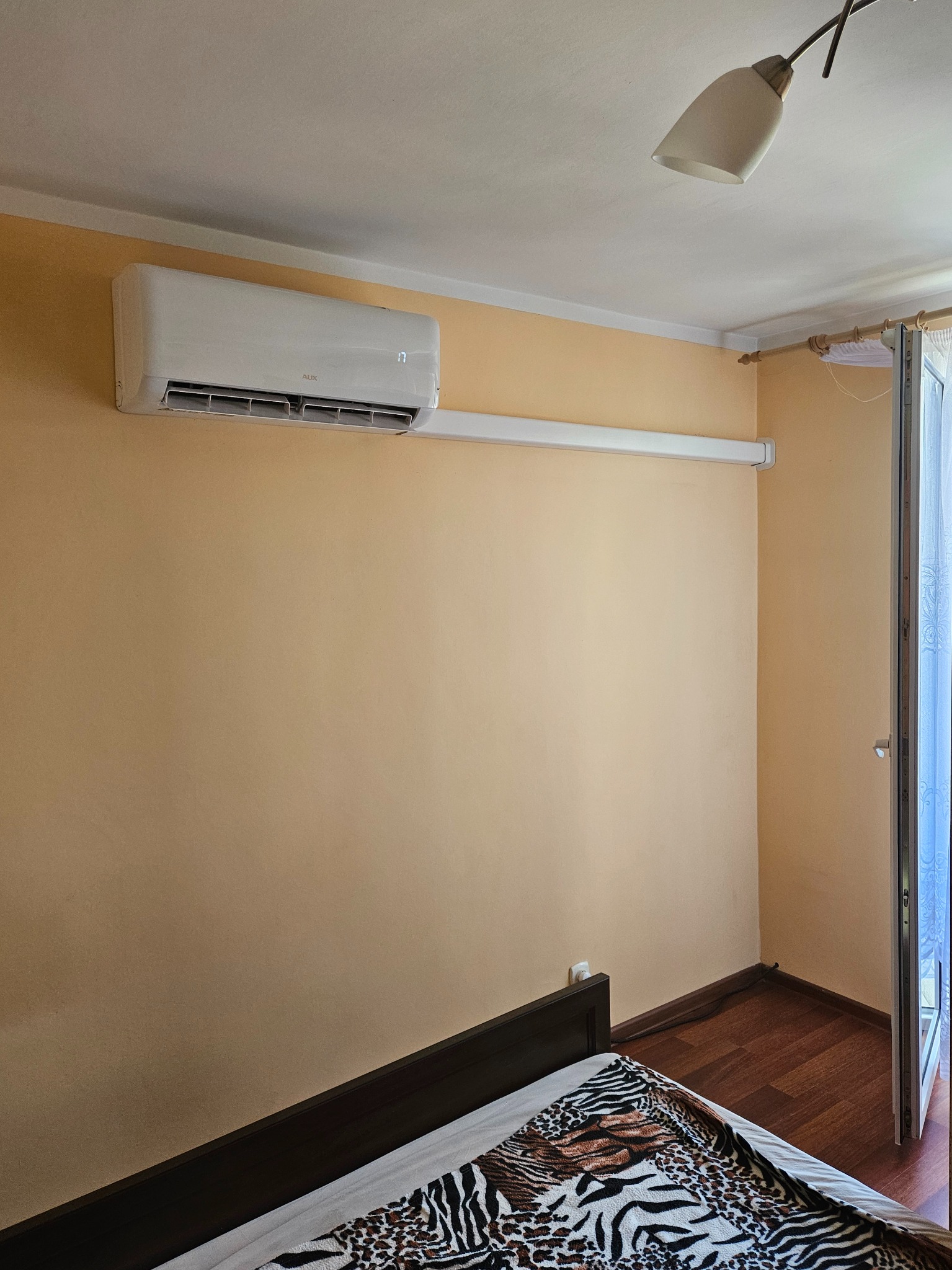 Biały klimatyzator AUX zamontowany na beżowej ścianie w sypialni, widoczny fragment łóżka z narzutą w zwierzęcy wzór, lampa sufitowa i otwarte drzwi balkonowe z firanką.