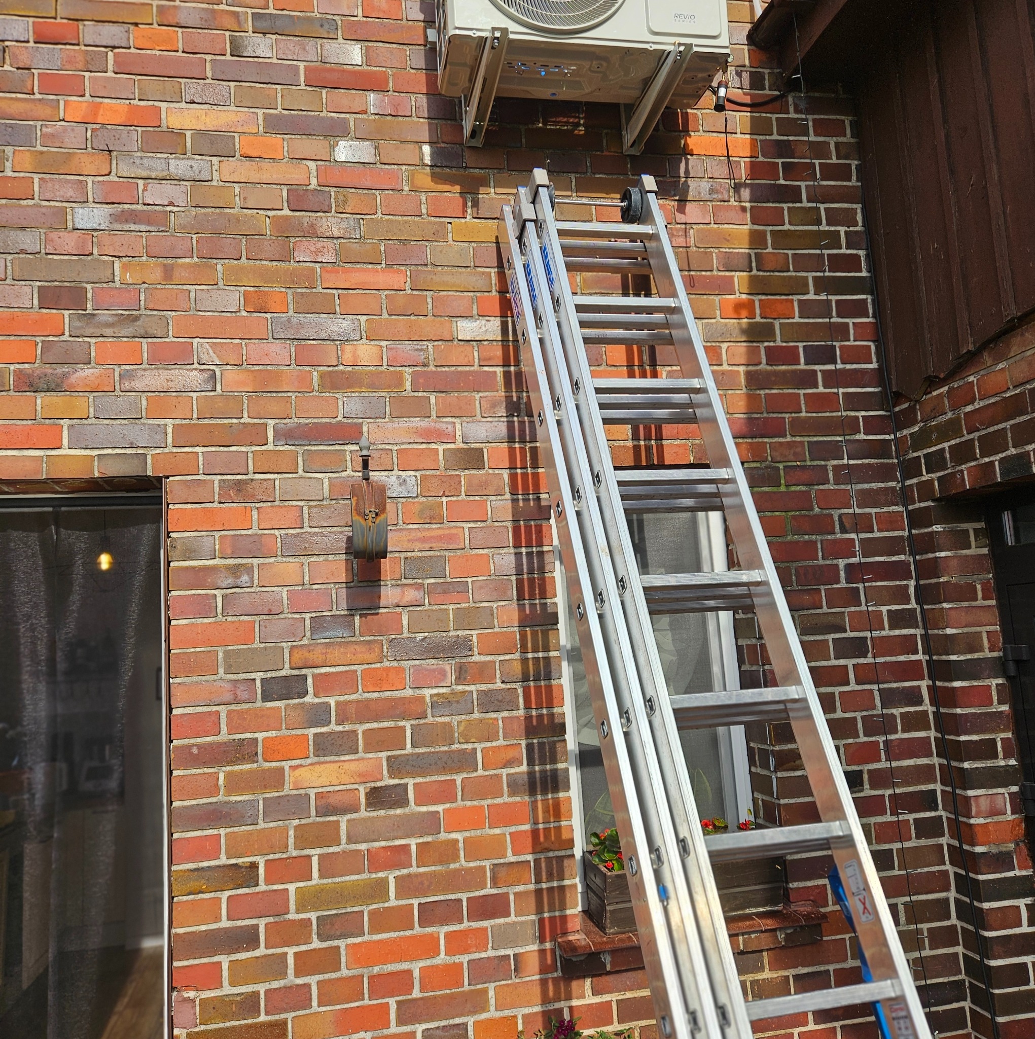 Zewnętrzna jednostka klimatyzacji zamontowana na ceglanej ścianie domu, z rozstawioną aluminiową drabiną opartą o ścianę.