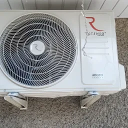 Biała jednostka zewnętrzna klimatyzacji Rotenso Imoto Series zamontowana na szarej, chropowatej ścianie, z fragmentem dekoracji świątecznej w tle.