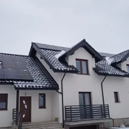 Biały dom jednorodzinny z ciemną dachówką i panelami słonecznymi na dachu, częściowo pokrytymi śniegiem, widok z dołu w pochmurny dzień.