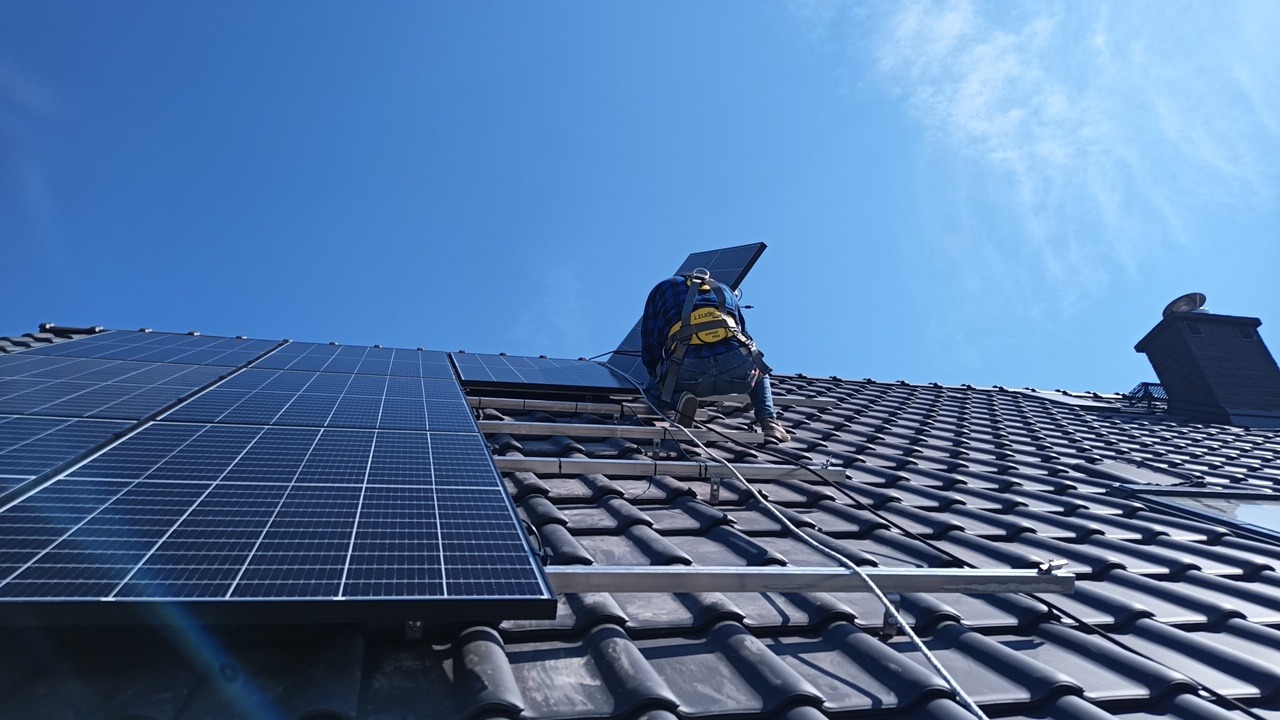 Montaż paneli słonecznych na dachu domu z czarną dachówką, pracownik w uprzęży zabezpieczającej, błękitne niebo w tle.