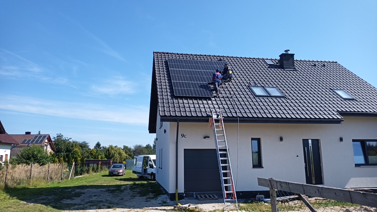 Montaż paneli słonecznych na dachu domu jednorodzinnego z czarną dachówką; dwóch monterów na drabinie, widoczny samochód dostawczy i fragment ogrodu.