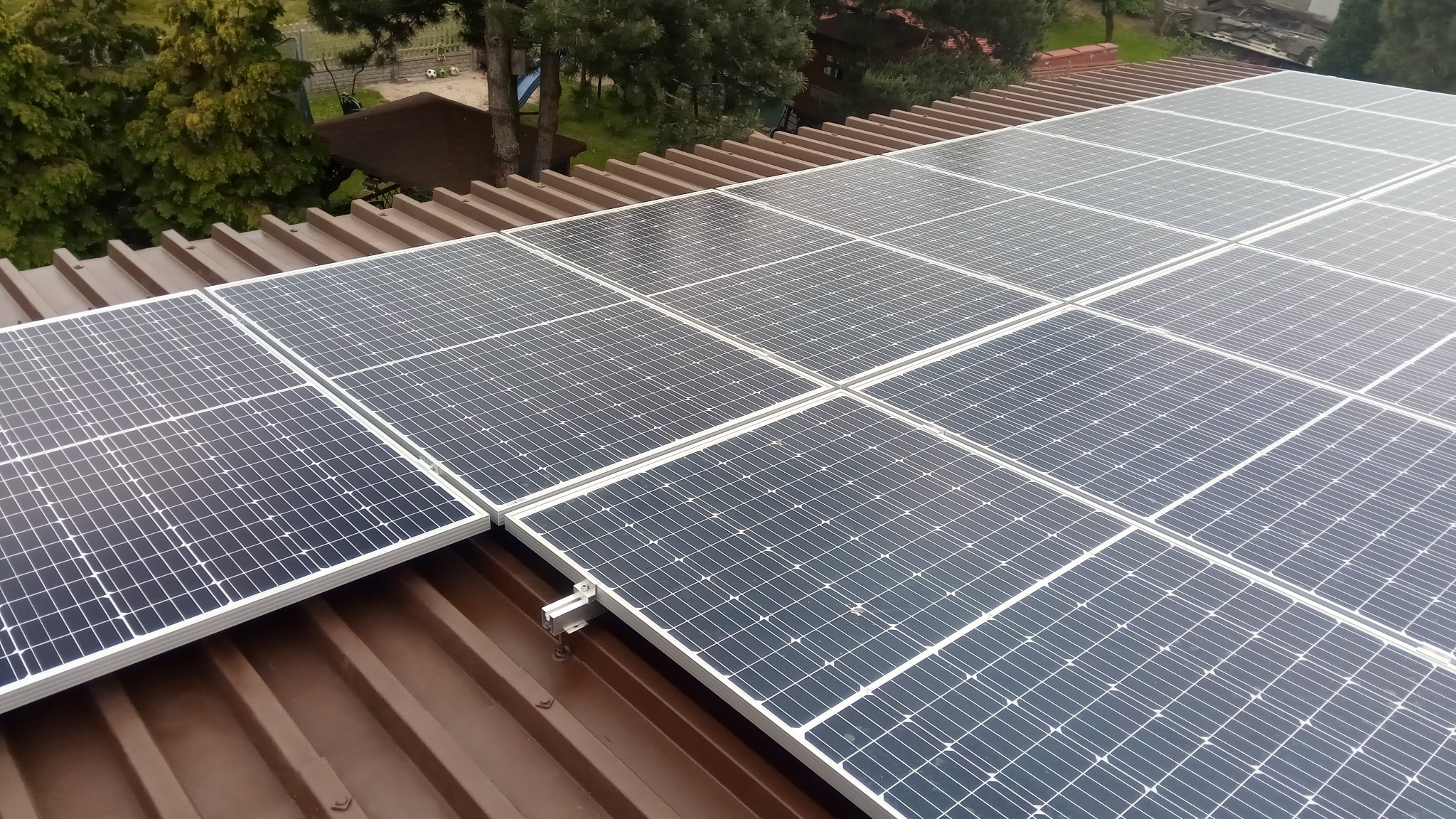 Panele słoneczne na brązowym, metalowym dachu z widocznymi elementami montażowymi, drzewa w tle.