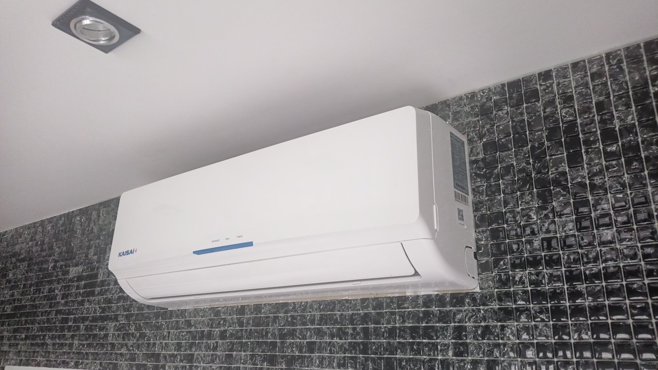 Biały klimatyzator marki Kaisai zamontowany na ścianie wyłożonej czarnymi, błyszczącymi płytkami mozaikowymi, widoczna również kwadratowa oprawa oświetleniowa w suficie.