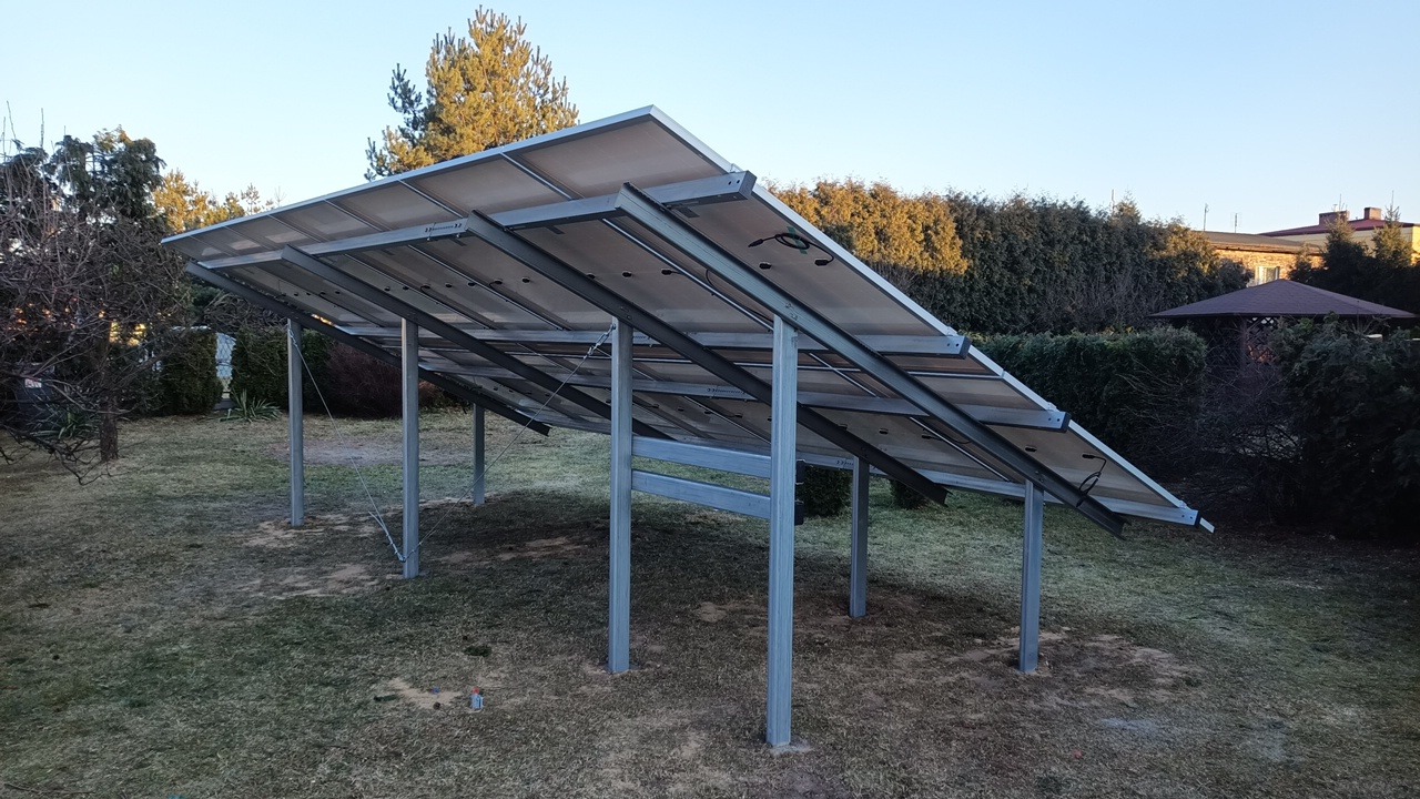 Widok od spodu na panele fotowoltaiczne zamontowane na metalowej konstrukcji na trawniku, w tle drzewa i altana.