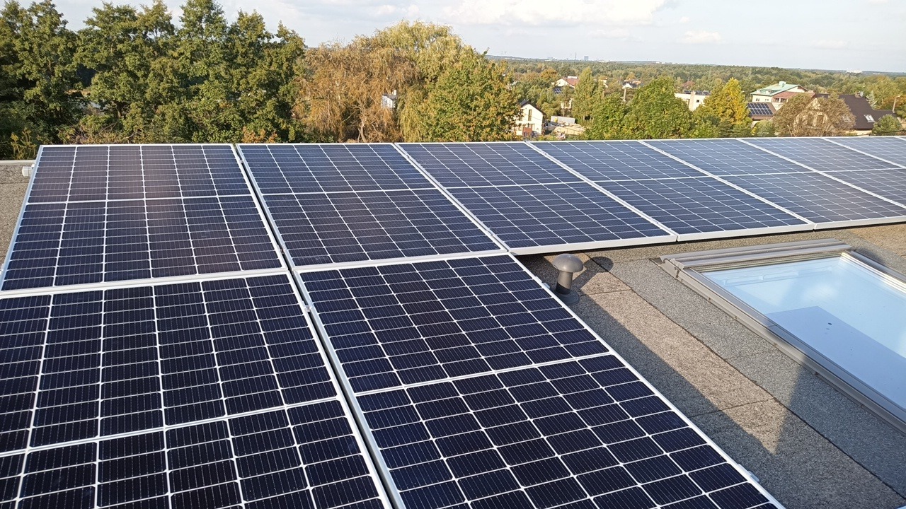 Panele fotowoltaiczne na płaskim dachu z widocznym świetlikiem dachowym i wentylacją, w tle drzewa i zabudowa mieszkalna.