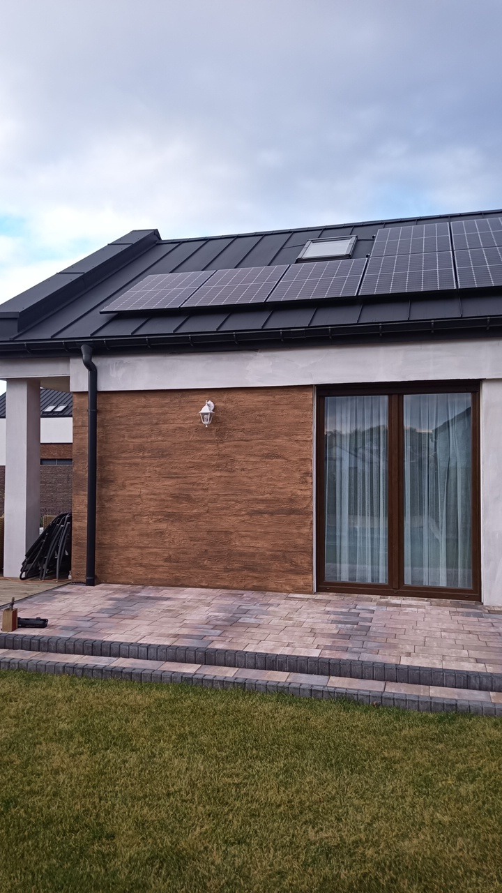 Nowoczesny dom jednorodzinny z panelami słonecznymi na dachu, widok z poziomu trawnika, z brązową elewacją imitującą drewno i dwuskrzydłowymi drzwiami tarasowymi.