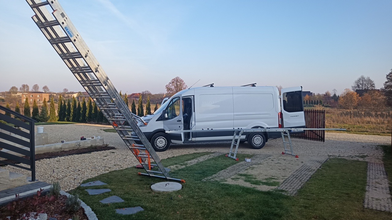 Biały samochód dostawczy z otwartymi drzwiami w Imielinie, obok rozstawiona długa drabina oparta o dach, mniejsza drabina przy tylnych drzwiach, teren pokryty żwirem.