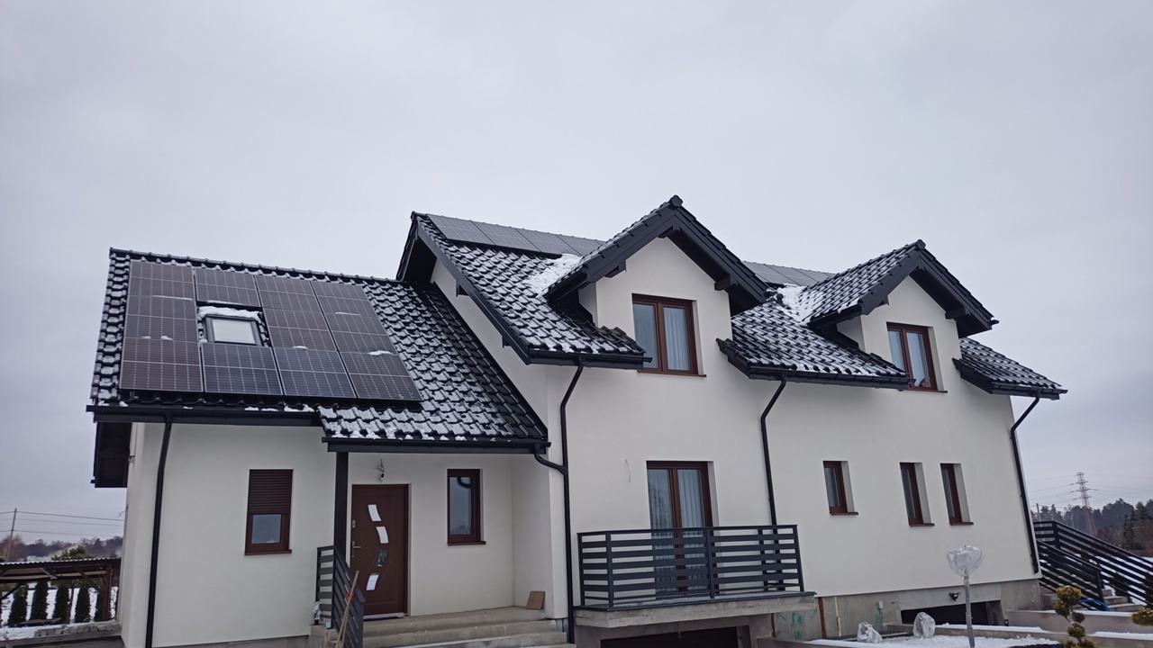 Biały dom jednorodzinny z ciemną dachówką i panelami słonecznymi na dachu, częściowo pokrytymi śniegiem, widok z dołu w pochmurny dzień.