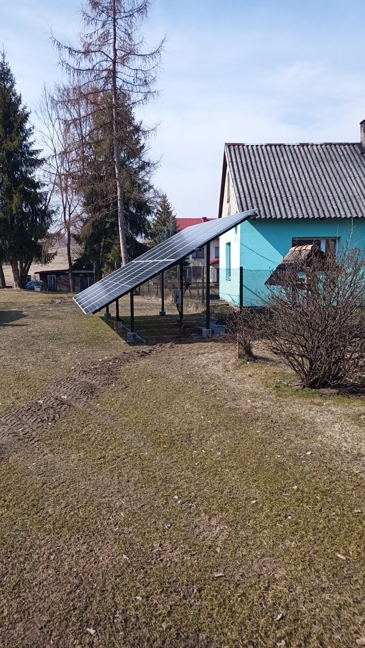 Panel słoneczny na metalowej konstrukcji w ogrodzie z widocznymi śladami opon, błękitny dom w tle.