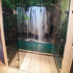 Szklana kabina prysznicowa z fototapetą wodospadu, chromowana armatura, płytki podłogowe w odcieniach beżu.