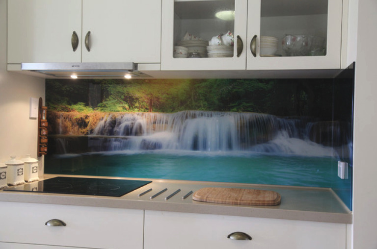 Szklany panel kuchenny z motywem wodospadu, wkomponowany między białe szafki, oświetlony punktowo lampkami podszafkowymi.