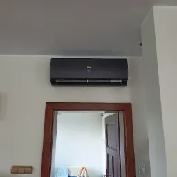 Czarna jednostka klimatyzacyjna Haier zamontowana na białej ścianie nad drewnianą futryną drzwiową, obok kwadratowy zegar ścienny.