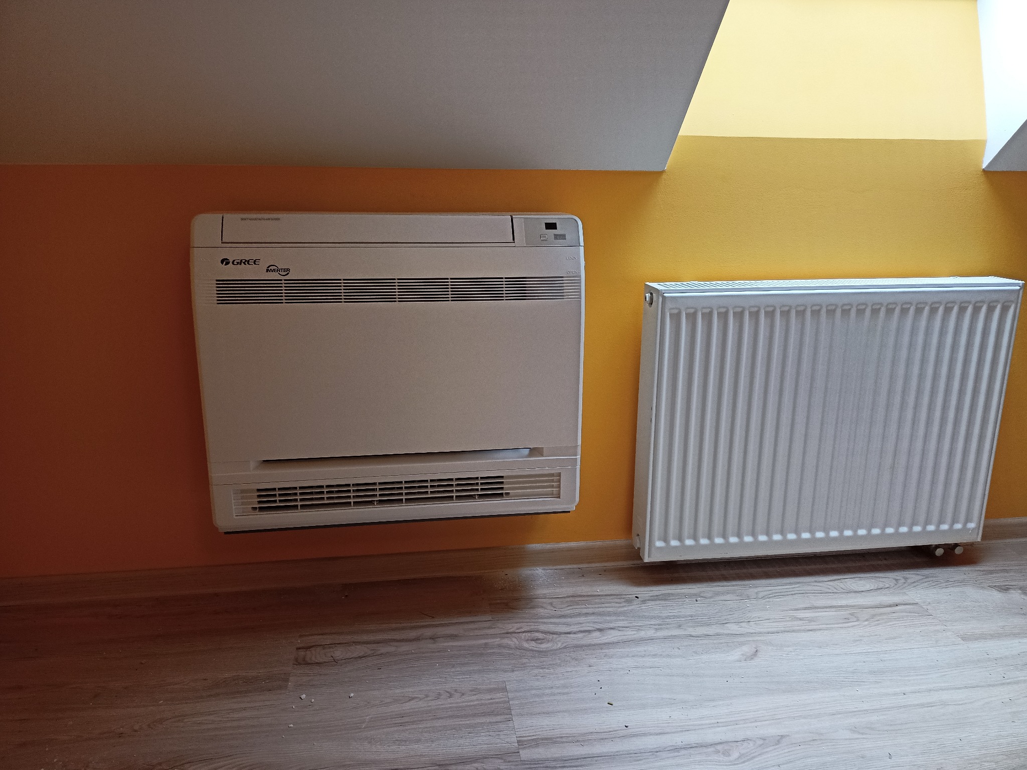 Biały klimatyzator podłogowy marki Gree zamontowany na pomarańczowej ścianie w pomieszczeniu ze skośnym sufitem, obok grzejnik żeberkowy.