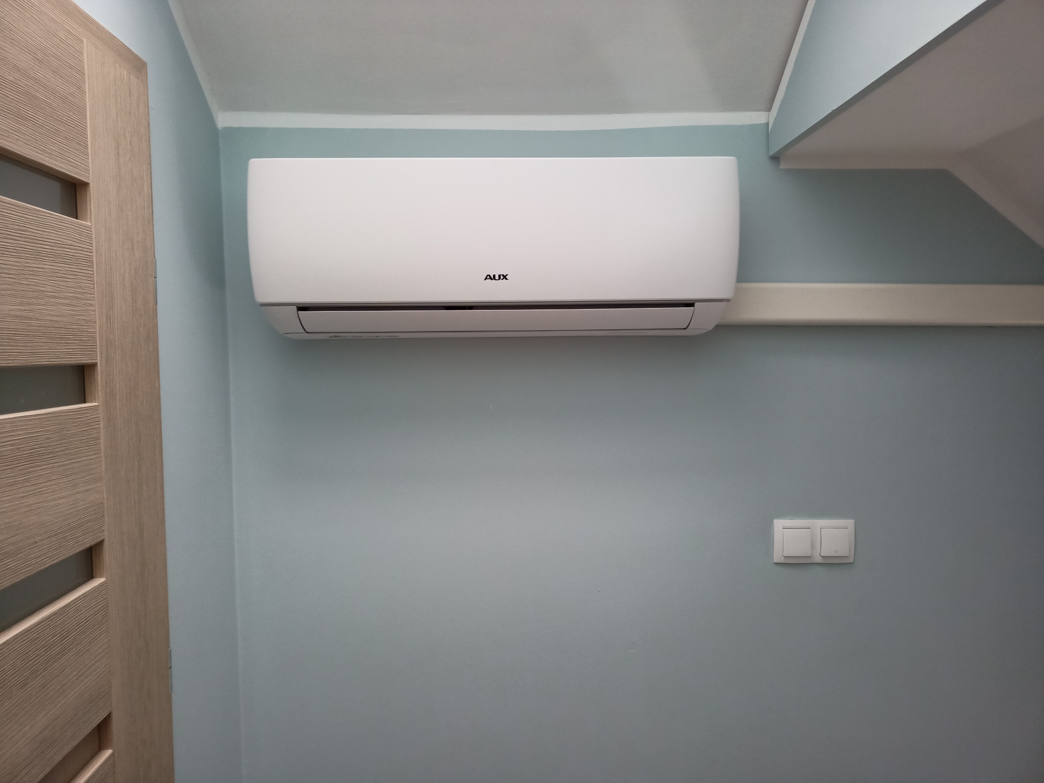 Biały klimatyzator marki AUX zamontowany na błękitnej ścianie w pomieszczeniu z drewnianymi drzwiami i włącznikiem światła.