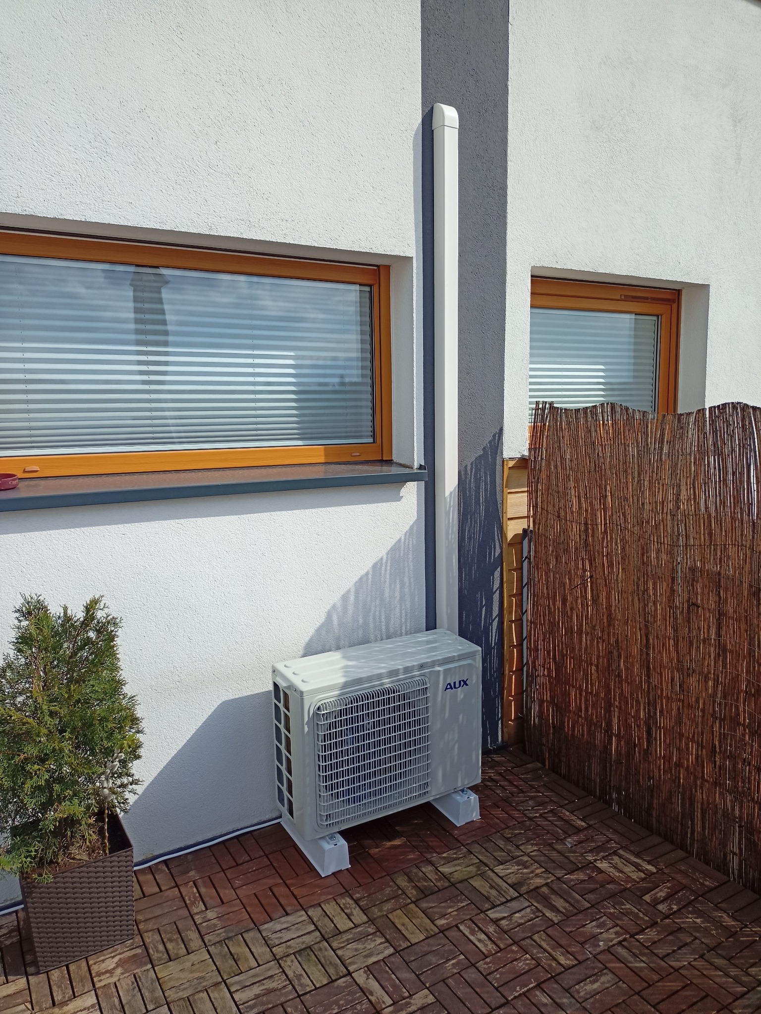 Zewnętrzna jednostka klimatyzacji AUX zamontowana na brązowej drewnianej podłodze tarasu przy ścianie budynku z dwoma oknami z żaluzjami, obok tui w donicy i bambusowej osłony.