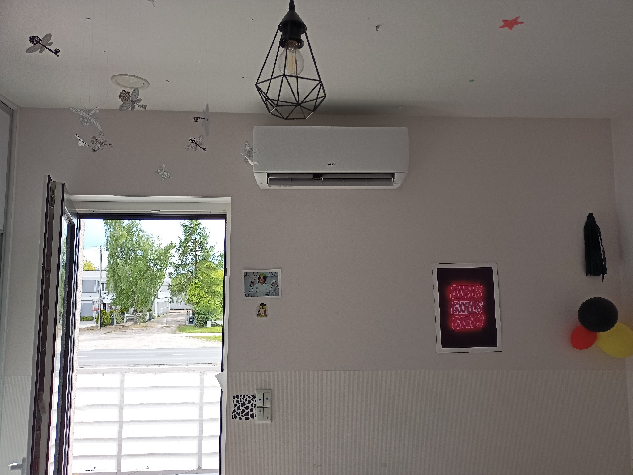 Wnętrze pokoju z zamontowaną białą klimatyzacją AUX, ozdobami sufitowymi w kształcie motyli i kluczy, lampą wiszącą w geometrycznej oprawie, otwartymi drzwiami balkonowymi oraz dekoracjami na ścianie.