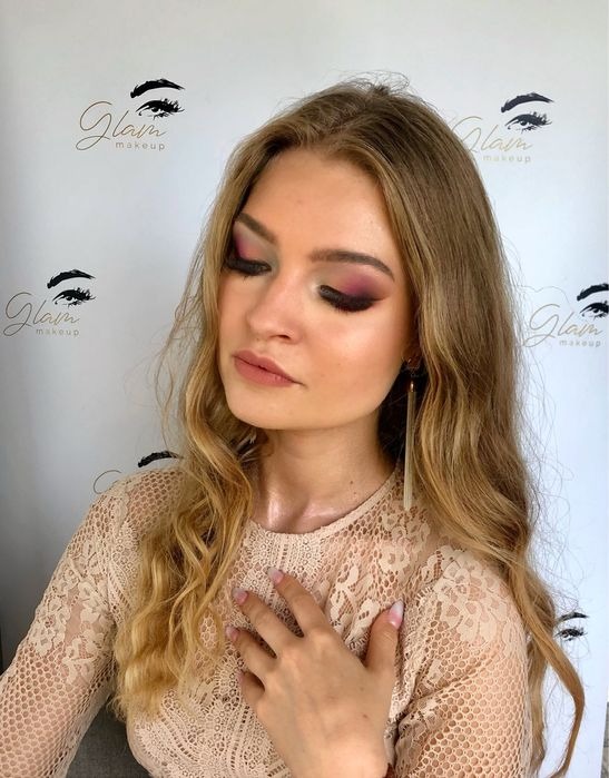 Portret kobiety z profesjonalnym makijażem oczu w odcieniach różu i zieleni, długie kolczyki i bluzka z koronki, dłoń na dekolcie, w tle logo salonu Glam makeup.