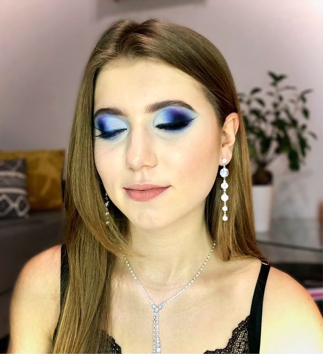 Portret kobiety z zamkniętymi oczami, prezentującej intensywny niebieski make-up oczu z ostrym, czarnym wykończeniem, długie, wiszące, srebrne kolczyki i pasujący naszyjnik z kryształkami, na tle...