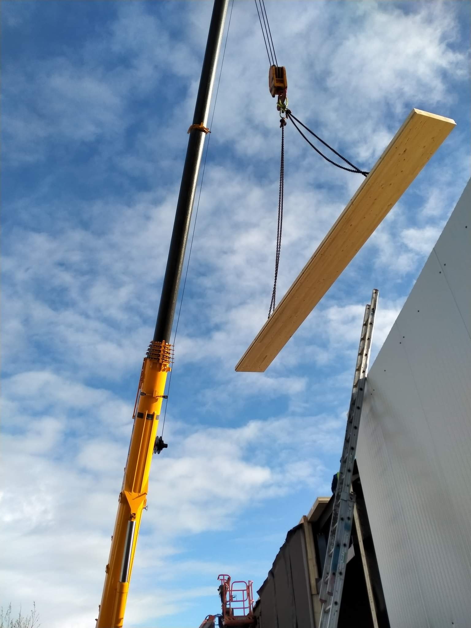 Żółty dźwig podnosi płytę warstwową na tle błękitnego nieba z chmurami, obok widoczna drabina oparta o ścianę budynku.