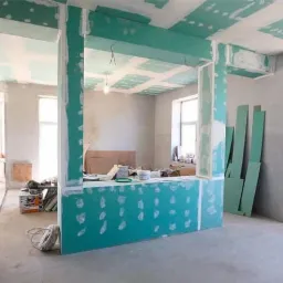 Wnętrze w trakcie remontu: płyty karton-gips zamontowane na ścianach i suficie, widoczne łączenia wymagające szpachlowania, surowe ściany, okna, narzędzia i materiały budowlane w tle.