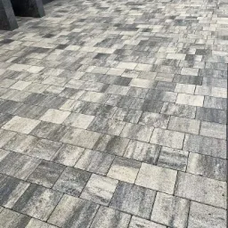 Ułożona kostka brukowa w odcieniach szarości i beżu, tworząca wzór na podjeździe, z ciemnoszarą elewacją budynku w tle.
