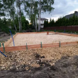 Przygotowany teren pod fundamenty domu jednorodzinnego z drewnianym szalunkiem, wypełniony jasnym żwirem. W tle widoczne drzewa i dom.