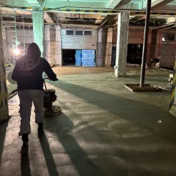 Pracownik w bluzie z logo firmy szlifuje świeżo wylaną, jasnoszarą posadzkę w hali przemysłowej. W tle widoczne materiały budowlane i brama garażowa.