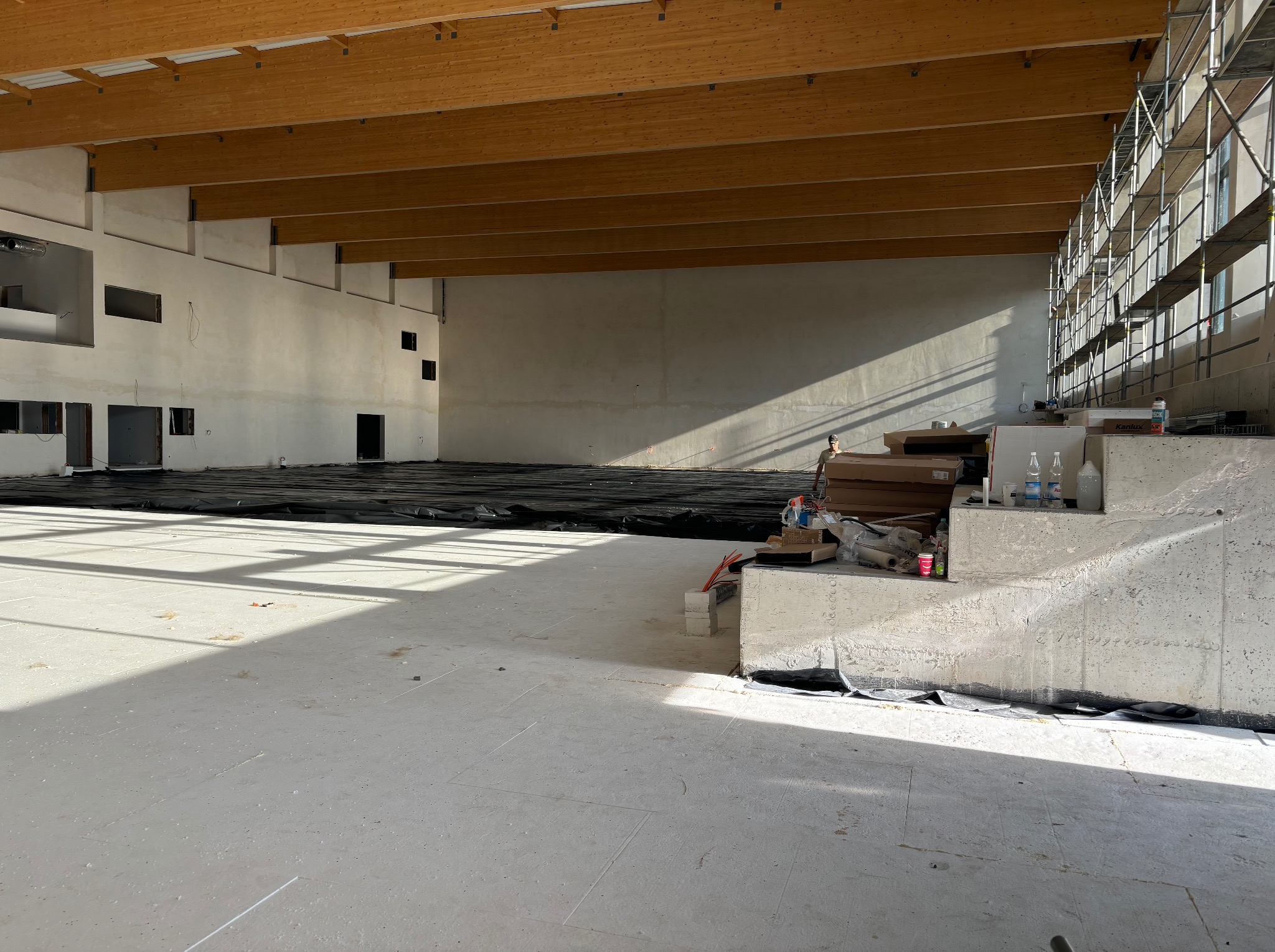 Wnętrze hali sportowej w trakcie prac przygotowawczych pod wylewkę posadzki przemysłowej, widoczne folie ochronne, narzędzia i materiały budowlane przy ścianie, drewniany strop i rusztowania przy...