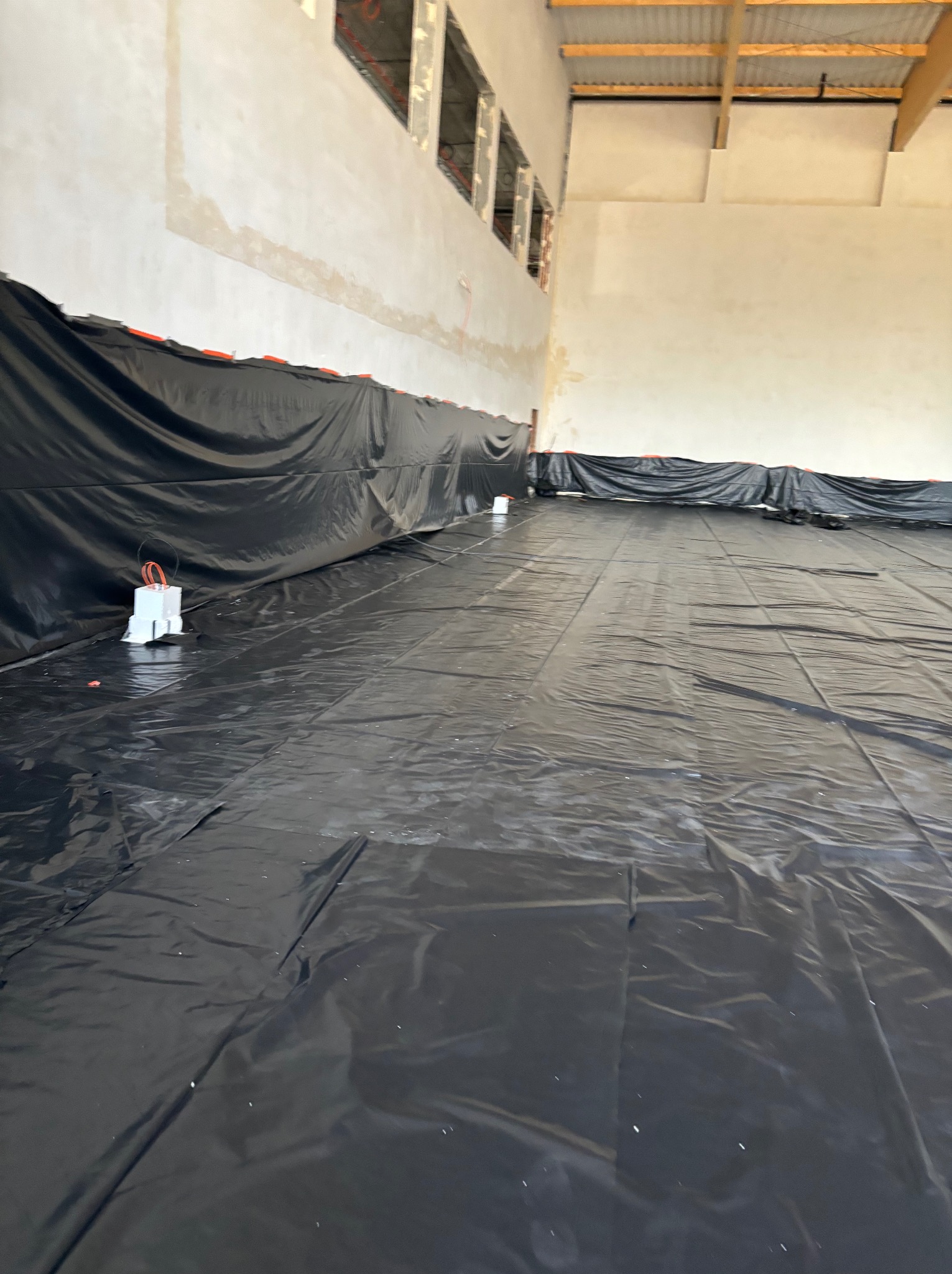 Przygotowanie podłoża pod wylewkę: rozłożona czarna folia ochronna z widocznymi przewodami elektrycznymi i zabezpieczonymi puszkami w dużym, surowym wnętrzu hali.