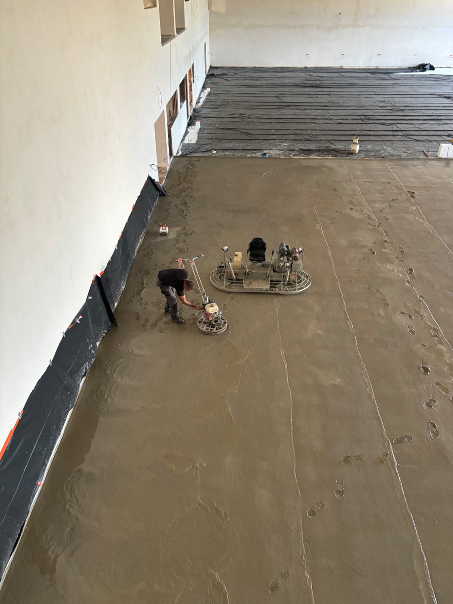 Pracownik w czarnej koszulce wyrównuje świeżo wylaną, brązową posadzkę za pomocą szlifierki do betonu; w tle widoczna druga szlifierka i czarna folia ochronna na ścianach.