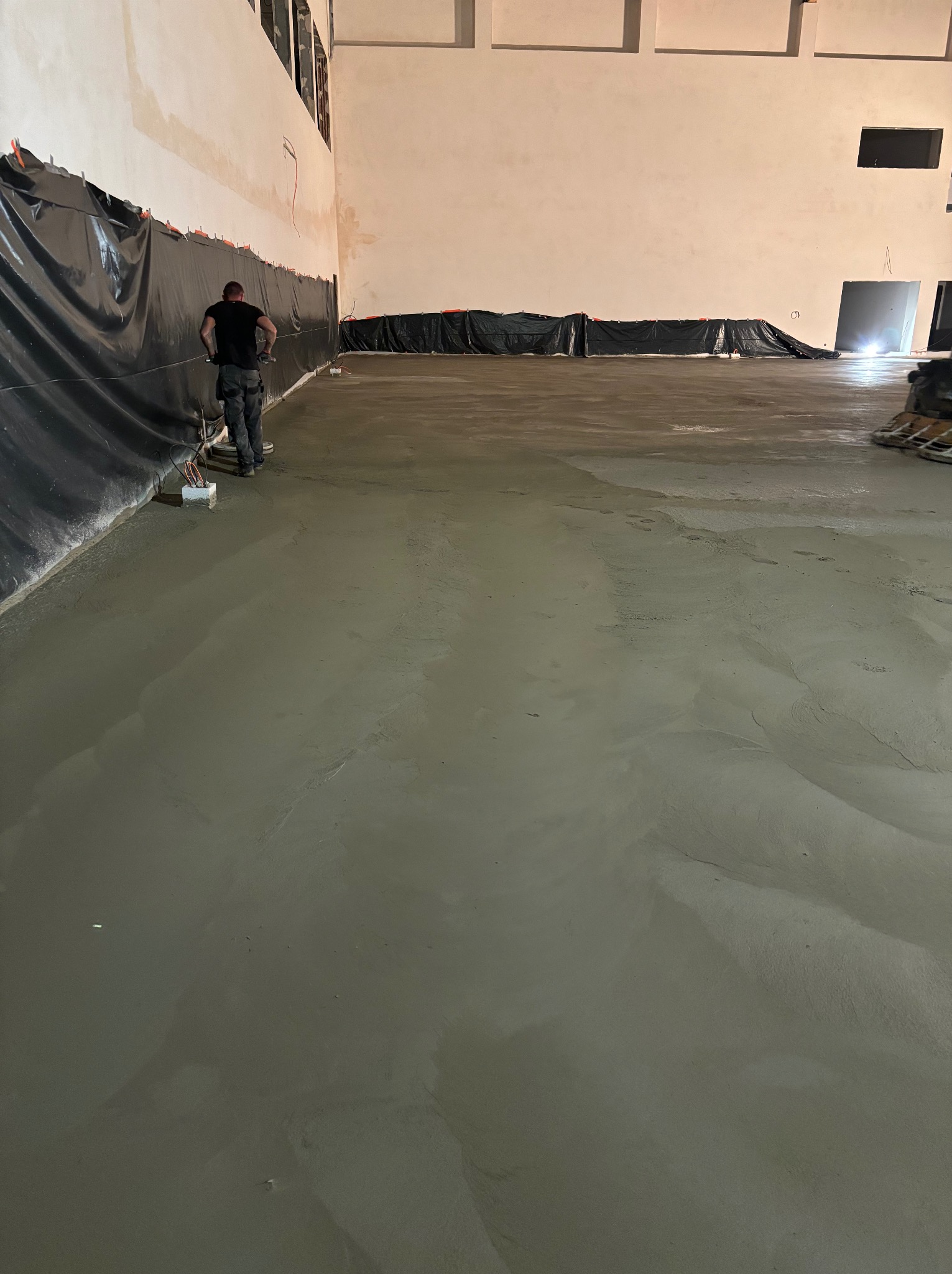 Aplikacja wylewki samopoziomującej w dużym, surowym pomieszczeniu, widoczny pracownik z narzędziami przy ścianie zabezpieczonej folią.