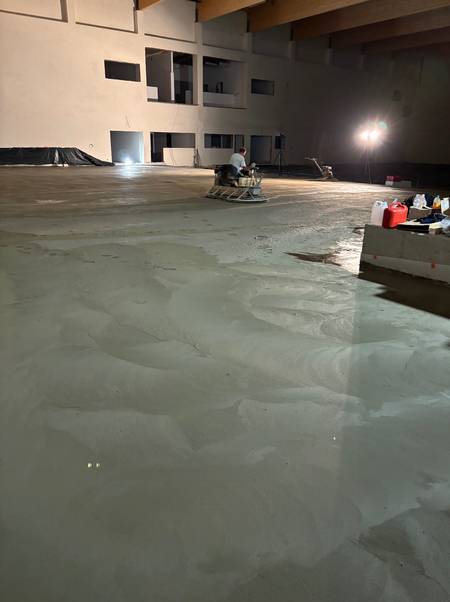 Pracownik wyrównuje świeżo wylaną posadzkę przemysłową za pomocą zacieraczki mechanicznej w przestronnym, niedokończonym wnętrzu hali. Widoczne ślady stóp na powierzchni, oświetlenie budowlane...