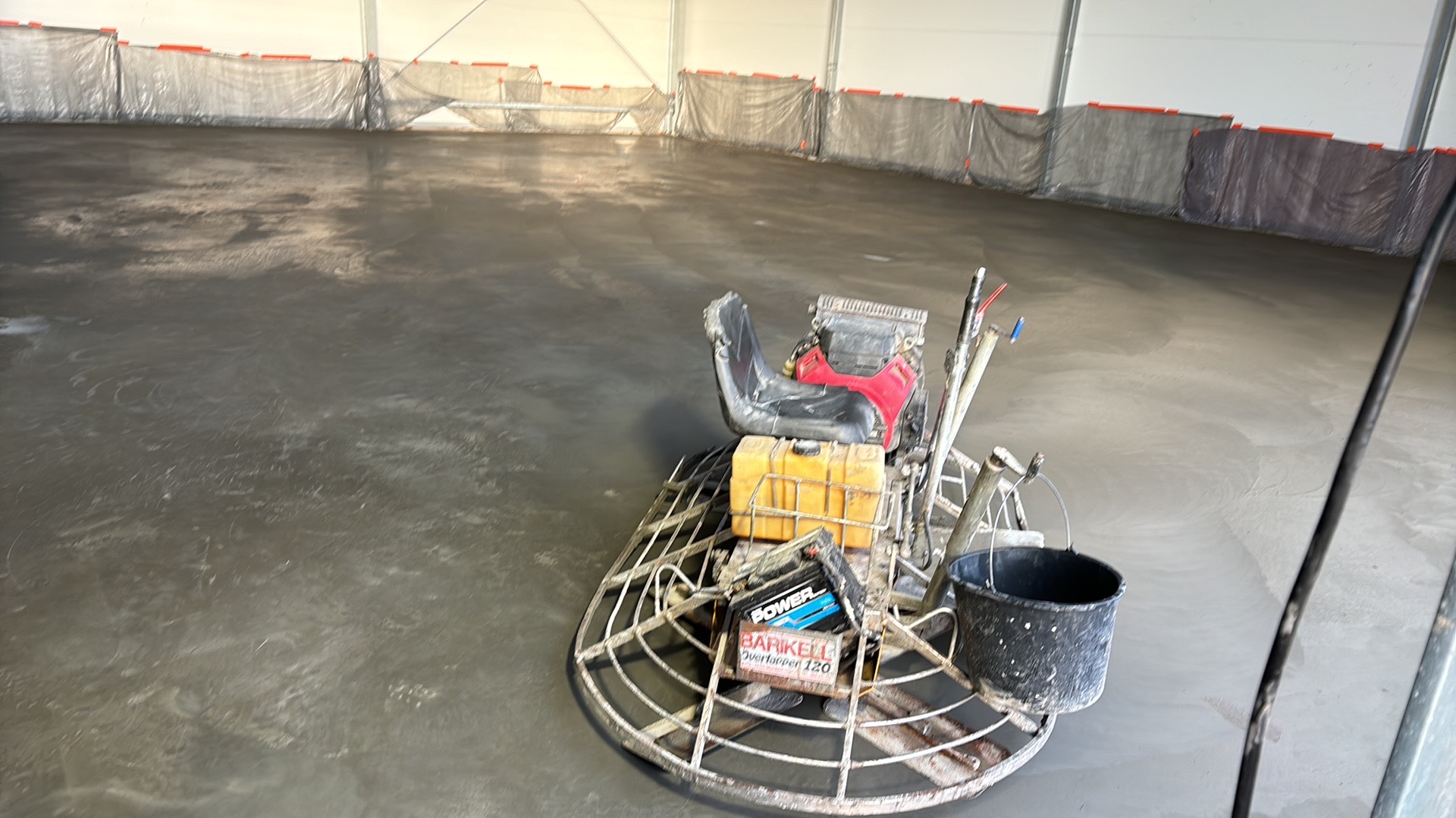 Świeżo wylana, szara posadzka w hali, zacierana mechaniczną zacieraczką Barikell Overlapper 120 z czerwonym siedziskiem i żółtym zbiornikiem paliwa, zabezpieczona folią na ścianach.