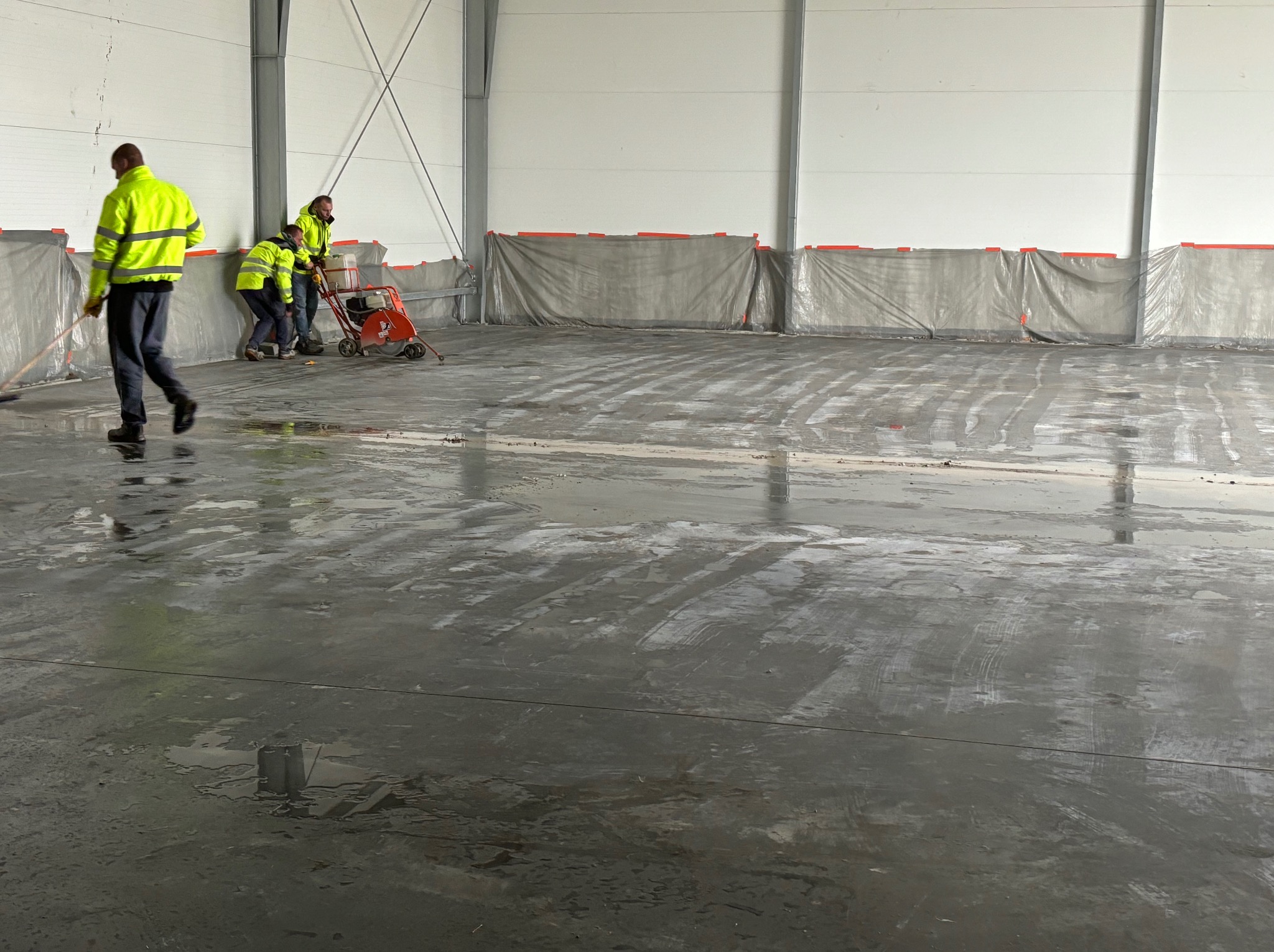Prace przygotowawcze do wylewki w hali: ekipa w jaskrawych kamizelkach szlifuje i zamiata betonową posadzkę, zabezpieczone ściany folią.