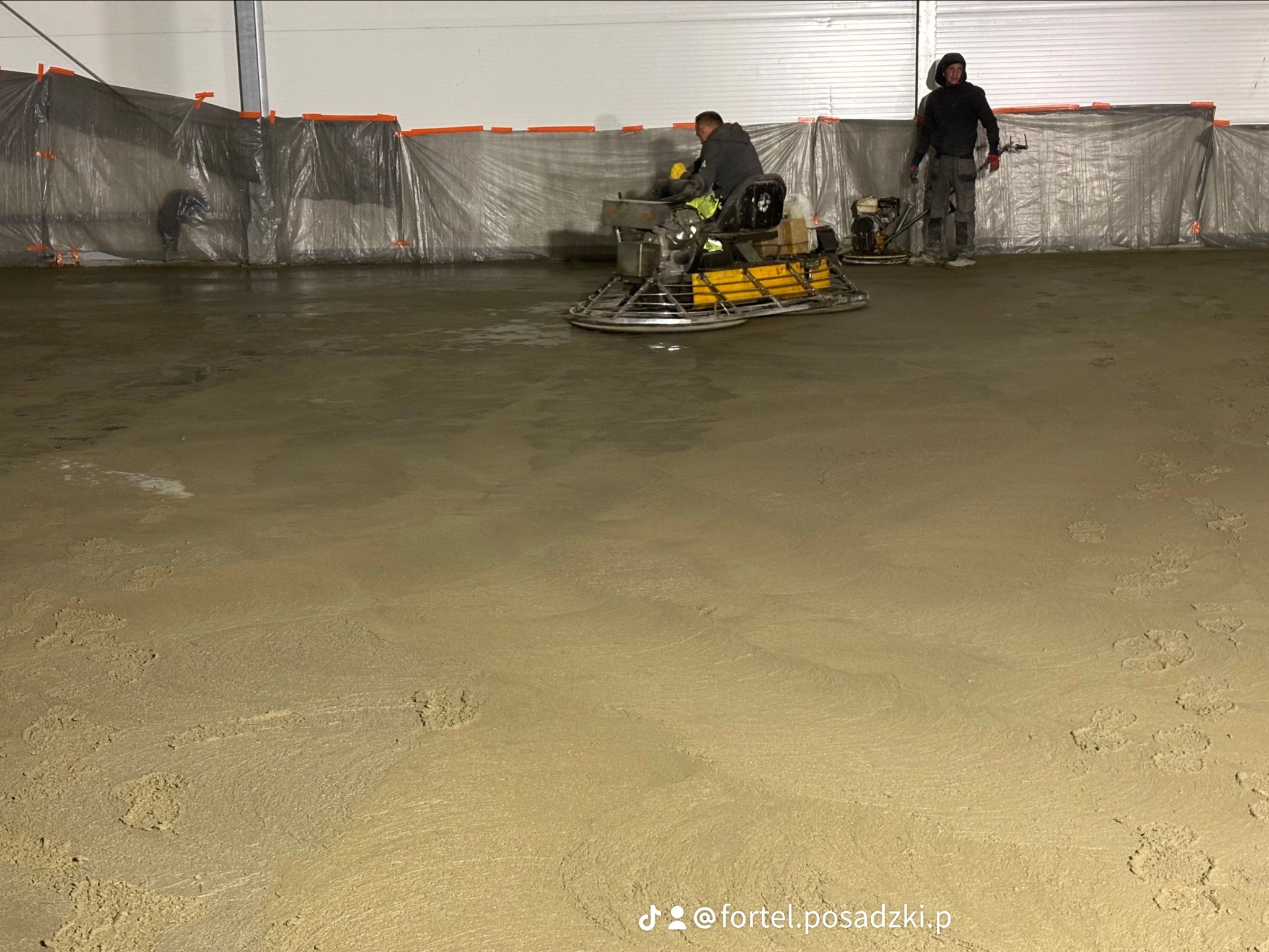 Pracownik wyrównuje świeżo wylaną, mokrą posadzkę przemysłową za pomocą zacieraczki mechanicznej w hali, drugi pracownik nadzoruje prace, widoczne ślady stóp na podłożu.
