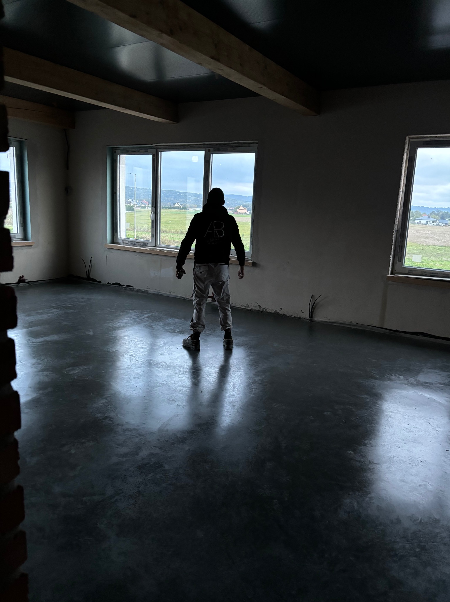 Wnętrze w stanie surowym z wylaną, ciemną posadzką, widok z tyłu na osobę w roboczym ubraniu stojącą na posadzce, okna z widokiem na krajobraz.