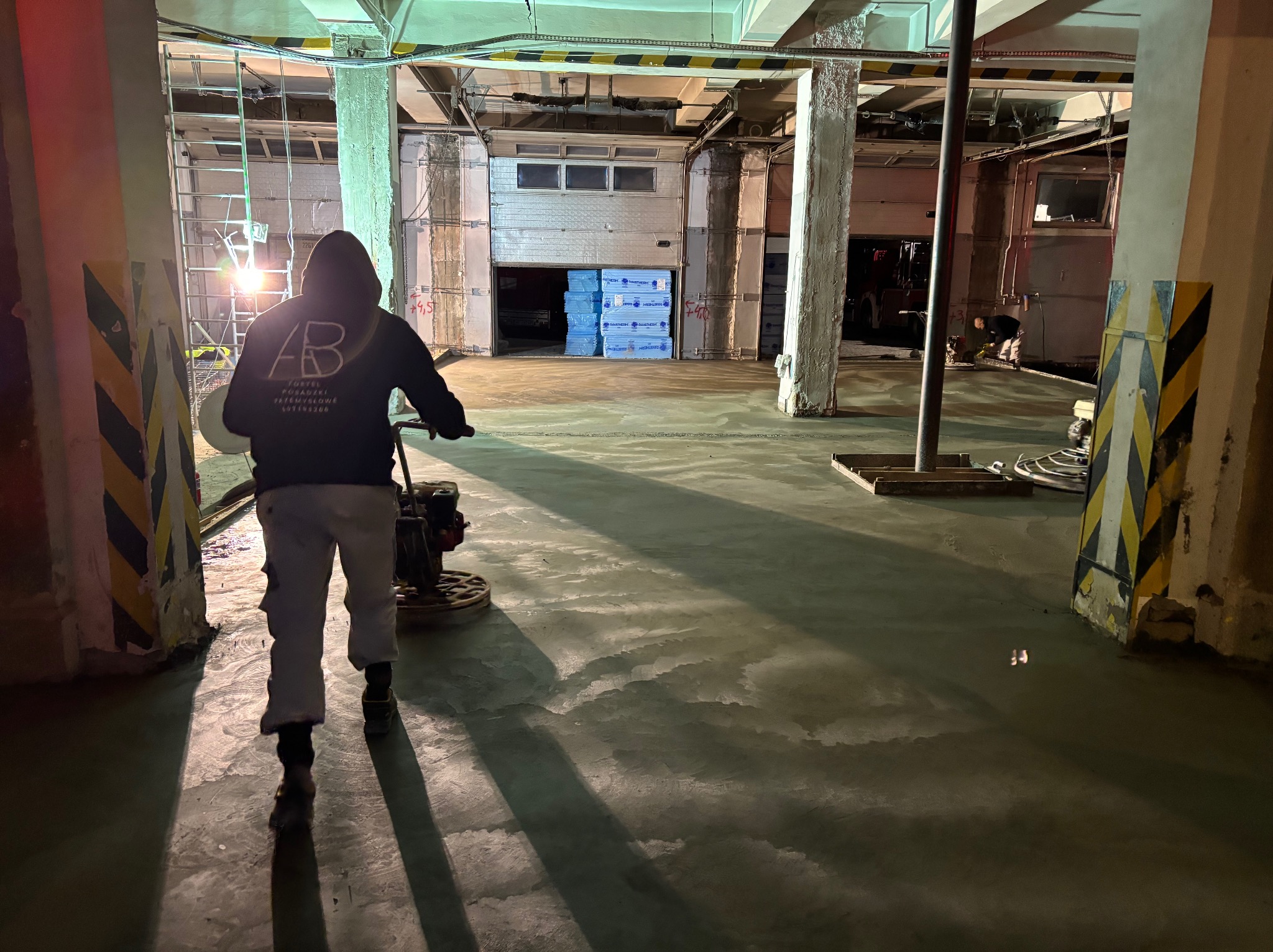 Pracownik w bluzie z logo firmy szlifuje świeżo wylaną, jasnoszarą posadzkę w hali przemysłowej. W tle widoczne materiały budowlane i brama garażowa.