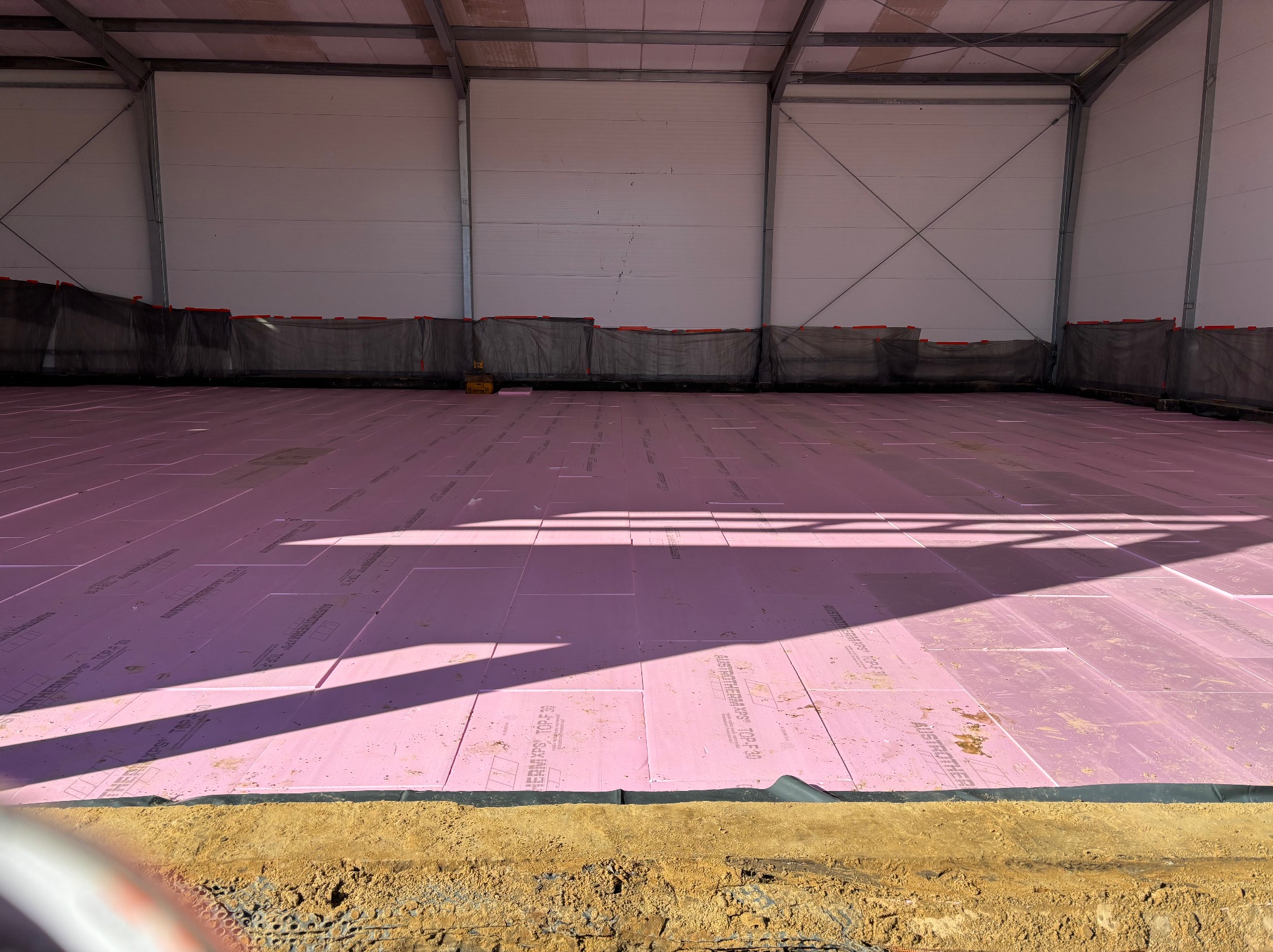 Przygotowanie podłoża pod posadzkę w hali przemysłowej: różowe płyty izolacyjne ułożone na czarnej folii, widoczny wykop z ziemią i fragment konstrukcji hali.