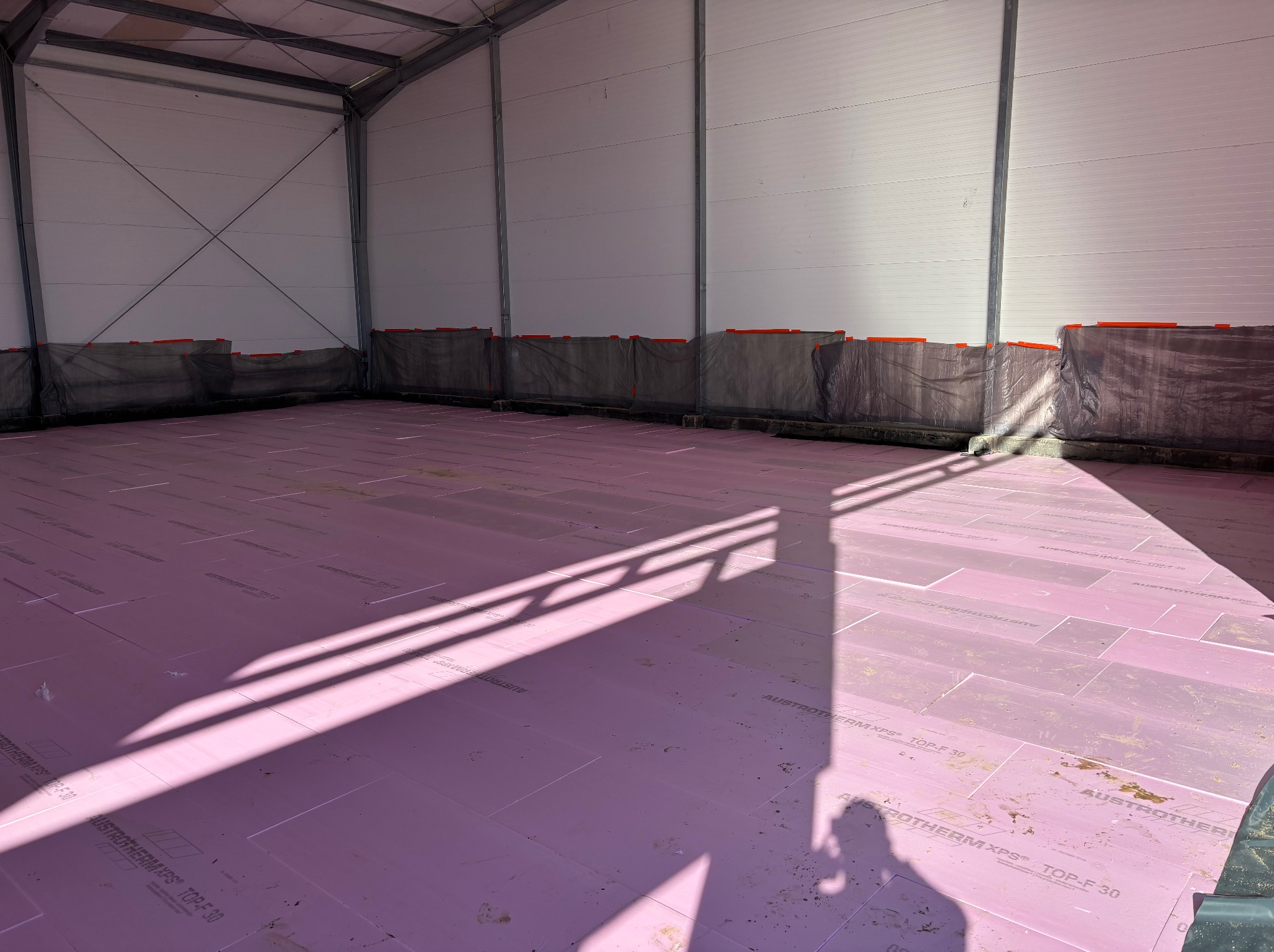 Przygotowanie podłoża w hali przemysłowej: różowe płyty izolacyjne Austrotherm XPS TOP-F 30 rozłożone na podłodze, cień konstrukcji dachu. Ściany hali wyłożone płytami warstwowymi, zabezpieczone...