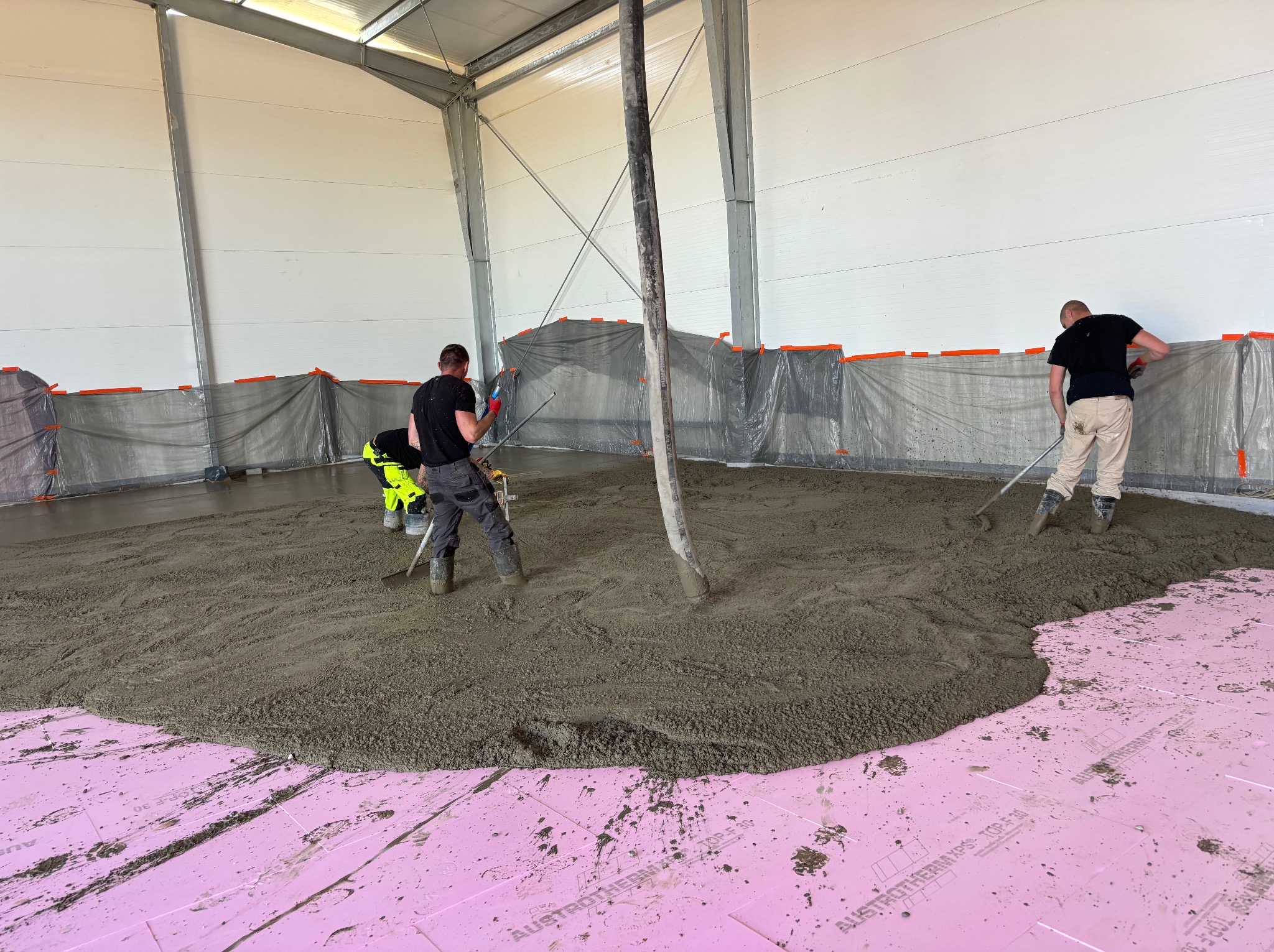 Wylewka posadzki w hali przemysłowej na warstwie izolacyjnej z różowych płyt styropianowych; widoczni trzej pracownicy w trakcie rozprowadzania betonu za pomocą narzędzi ręcznych.