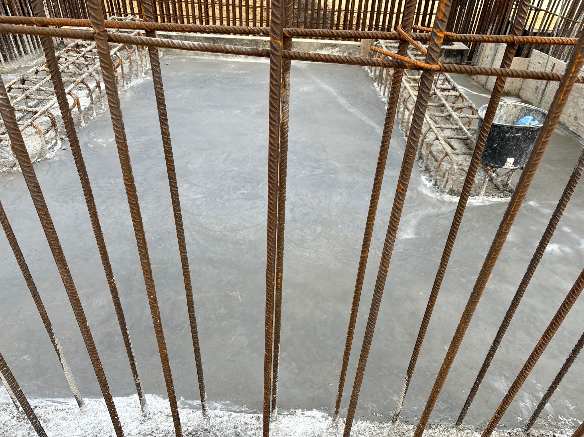 Świeżo wylana, szara betonowa posadzka przemysłowa, widoczna przez rustykalne, zardzewiałe pręty zbrojeniowe, z czarnym wiadrem w tle.