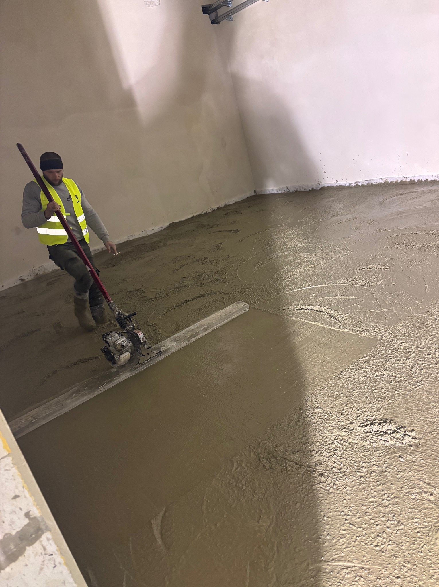 Pracownik w kamizelce odblaskowej wyrównuje wylewkę posadzki przemysłowej za pomocą listwy wibracyjnej w surowym wnętrzu.