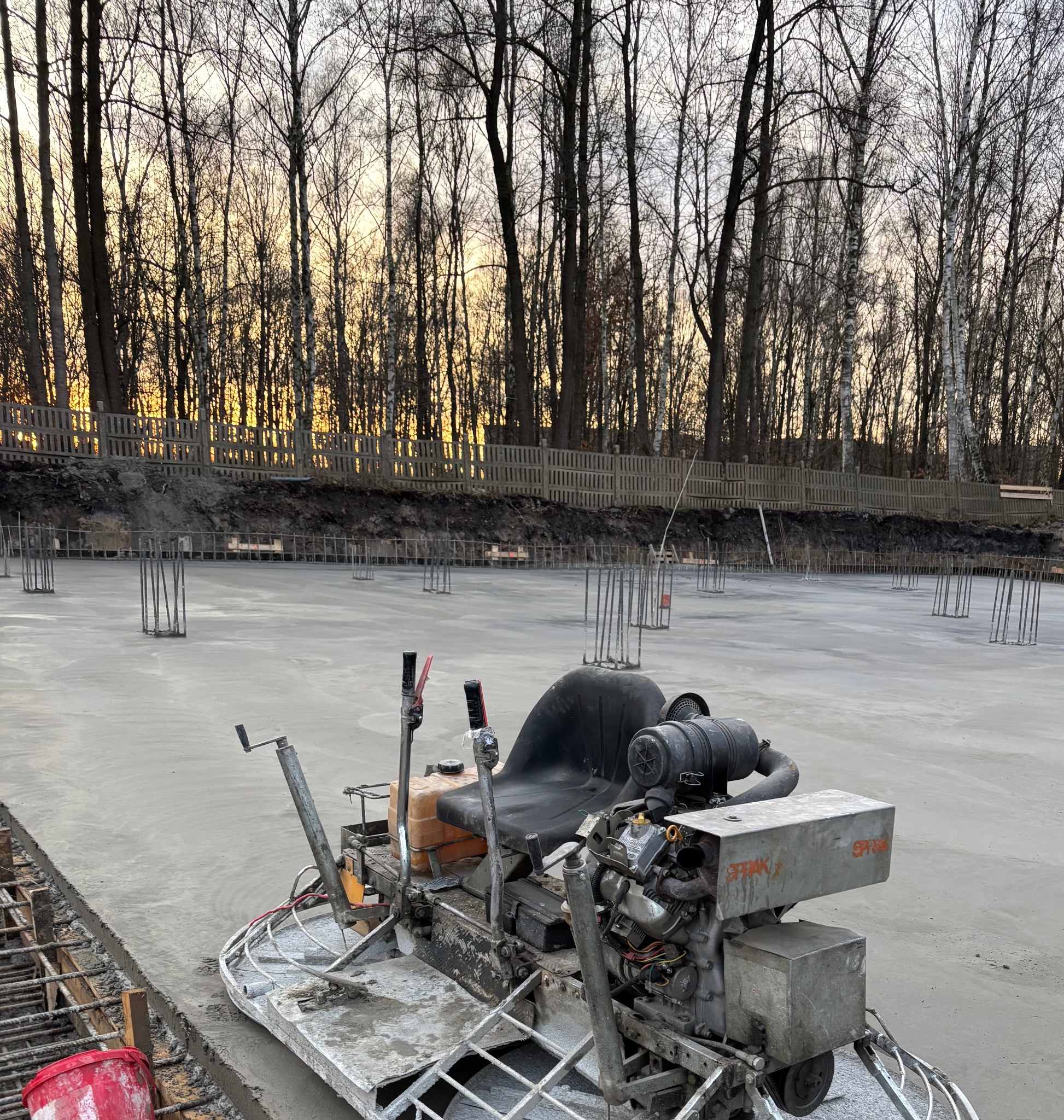 Wyrównywanie świeżo wylanej, szarej posadzki przemysłowej w Sosnowcu za pomocą maszyny z siedziskiem, widoczne wystające pręty zbrojeniowe i drzewa w tle o zmierzchu.