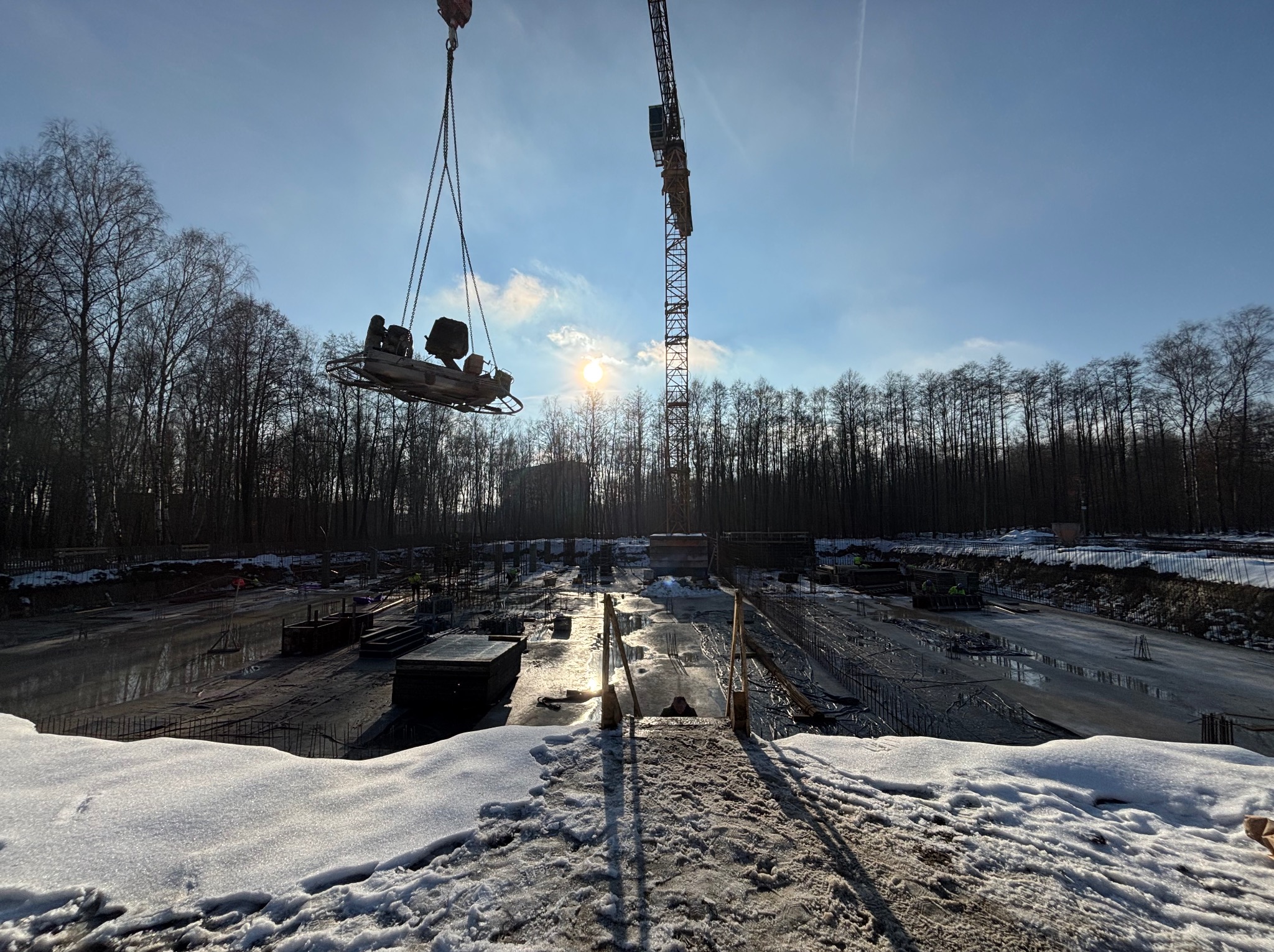 Przygotowanie podłoża pod wylewkę na placu budowy w zimowej scenerii, z widocznym dźwigiem podnoszącym ubijak wibracyjny i zbrojeniem w tle.
